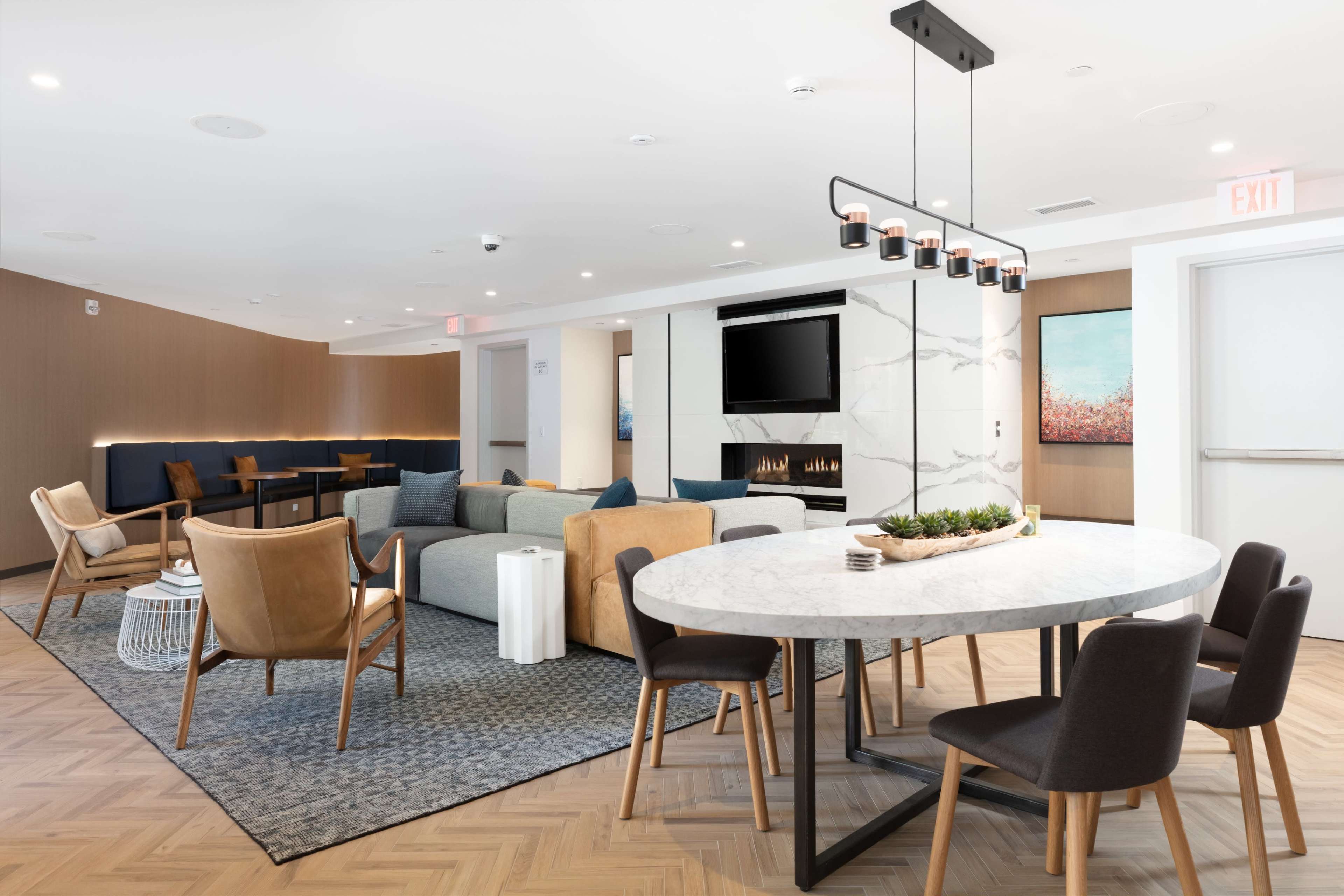 A modern lounge area featuring a circular marble dining table surrounded by various chairs, a sectional sofa, and an accent wall with a mounted television.