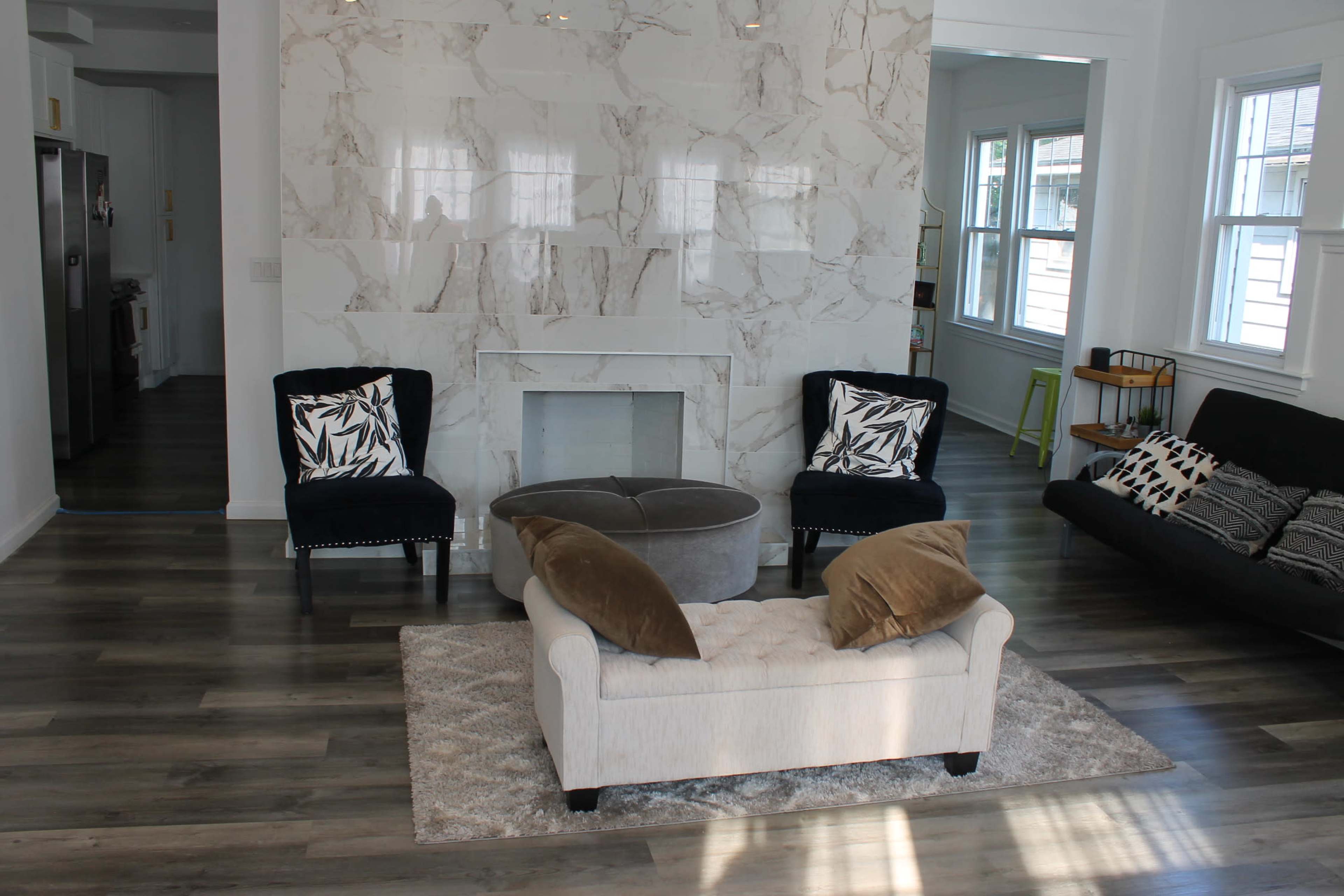 A spacious living area featuring a large decorative wall, two black armchairs, a white couch with pillows, and a round ottoman on a light rug.