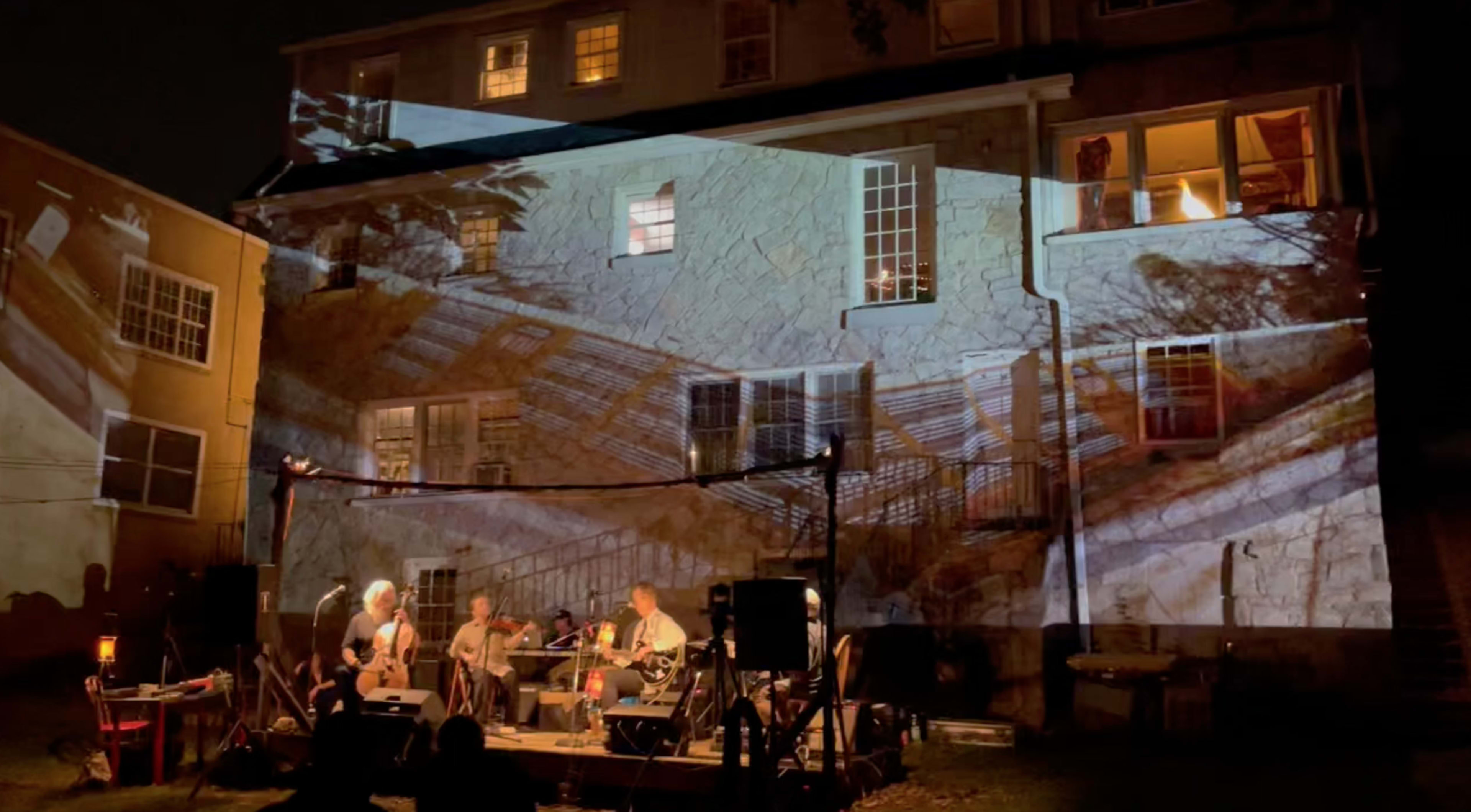 A band performs in front of a house with a large projection of its stone wall and windows on the building behind them.