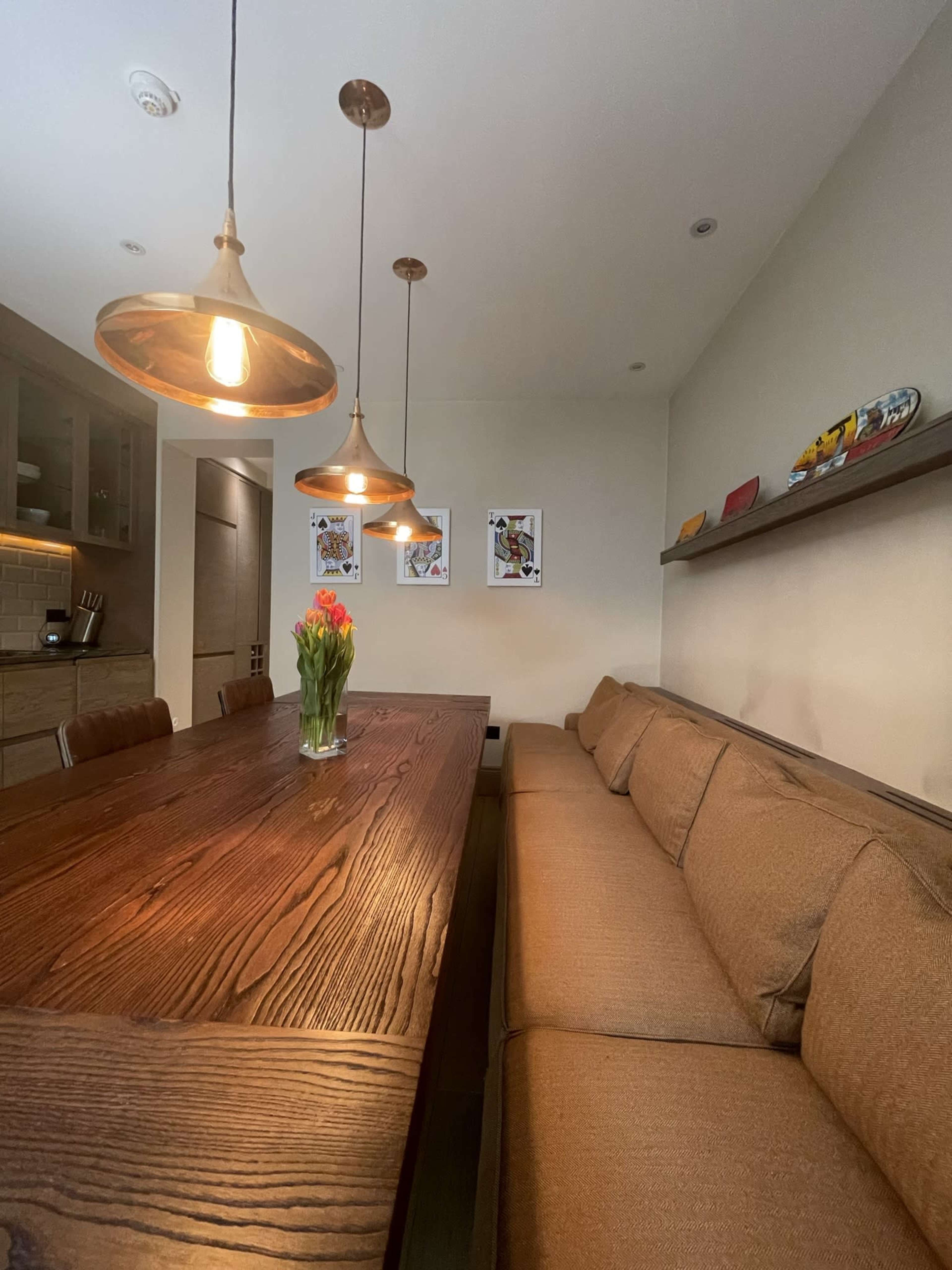 A long wooden dining table is accompanied by a brown upholstered bench, with pendant lights hanging above and artwork displayed on the wall.