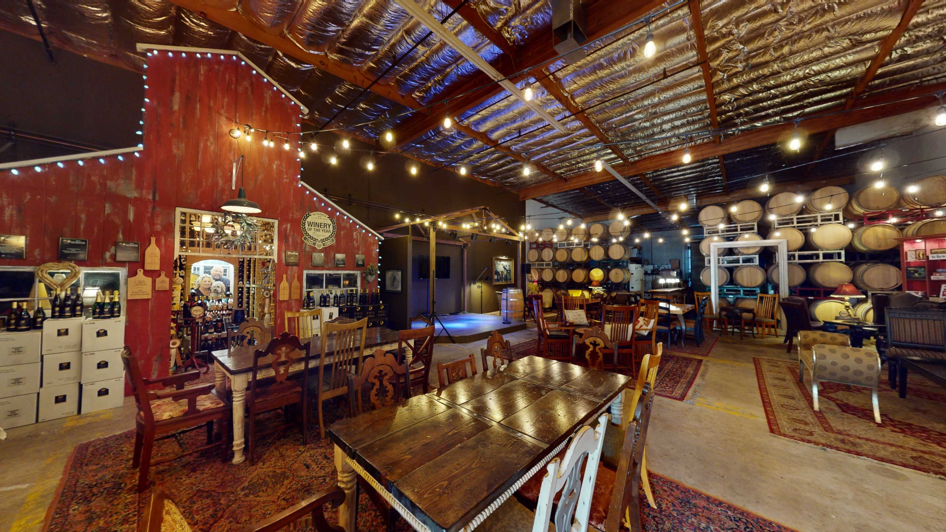 A rustic wine tasting room with wooden tables, a barn-style wall, and rows of wine barrels in the background, illuminated by string lights.