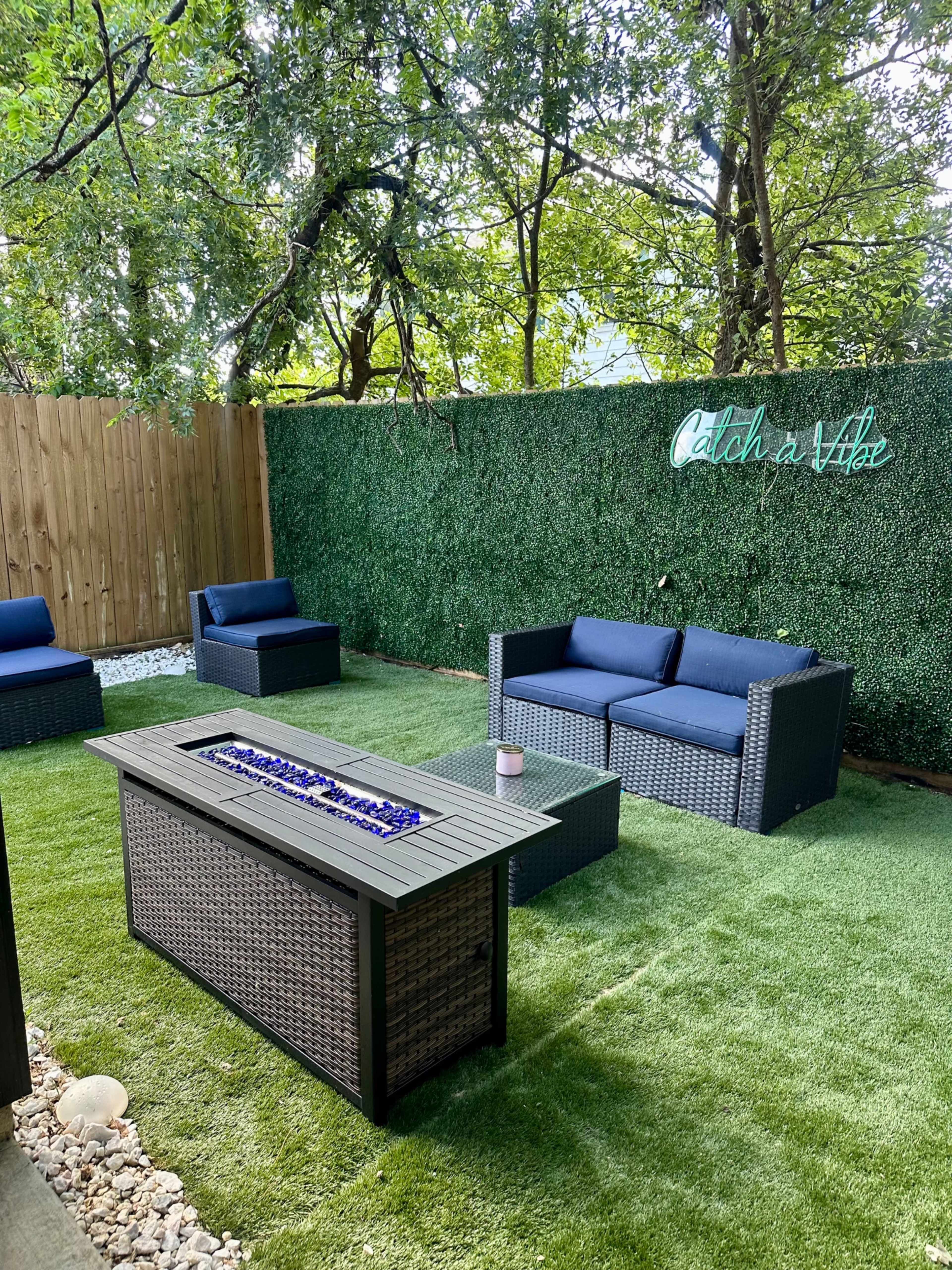 A landscaped outdoor seating area with two blue couches, a fire table with blue glass stones, and a green hedge backdrop featuring the phrase "Catch a Vibe."