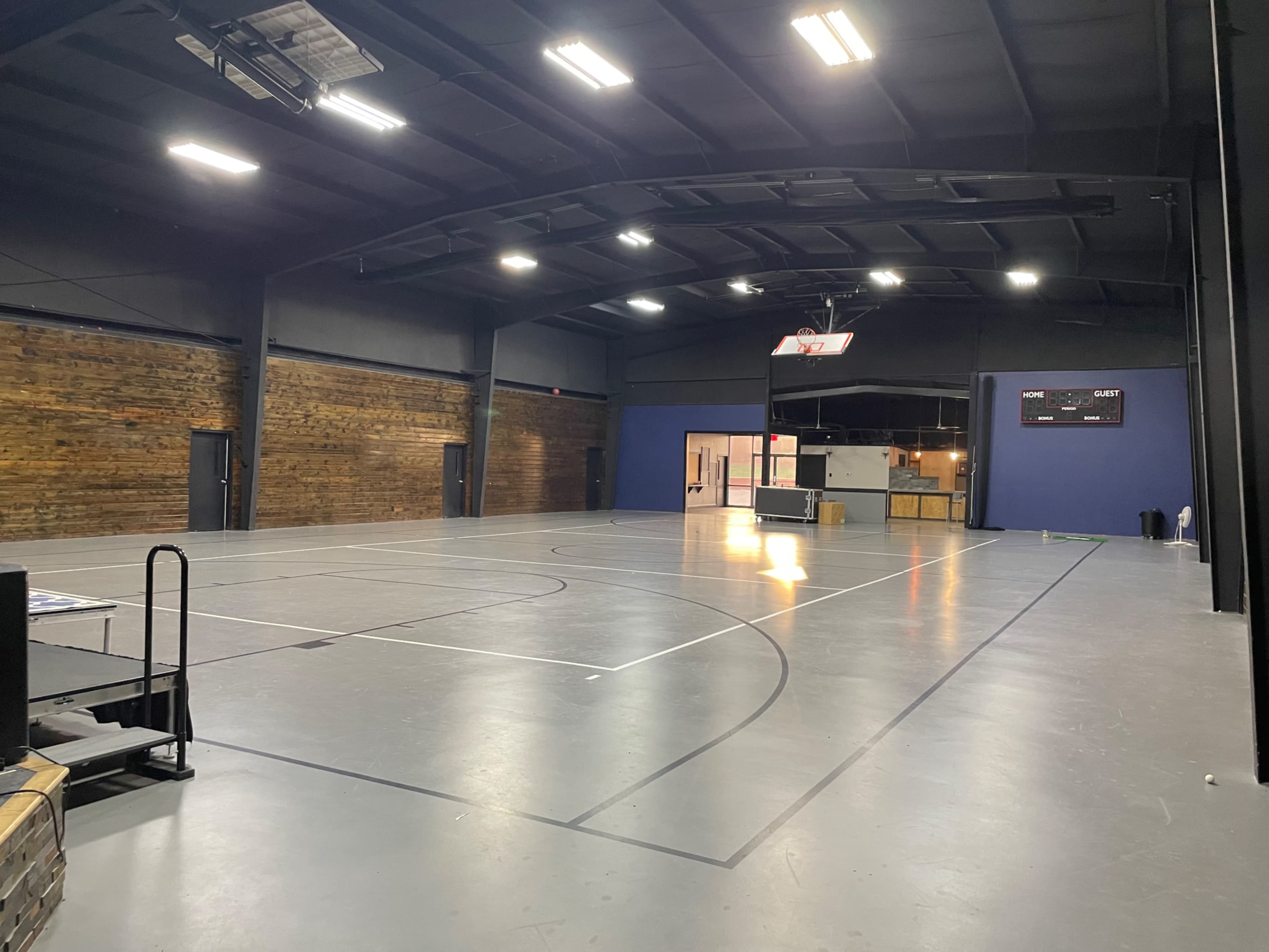 The image shows an empty indoor basketball court with a wooden wall, gray flooring, and a scoreboard in the background.
