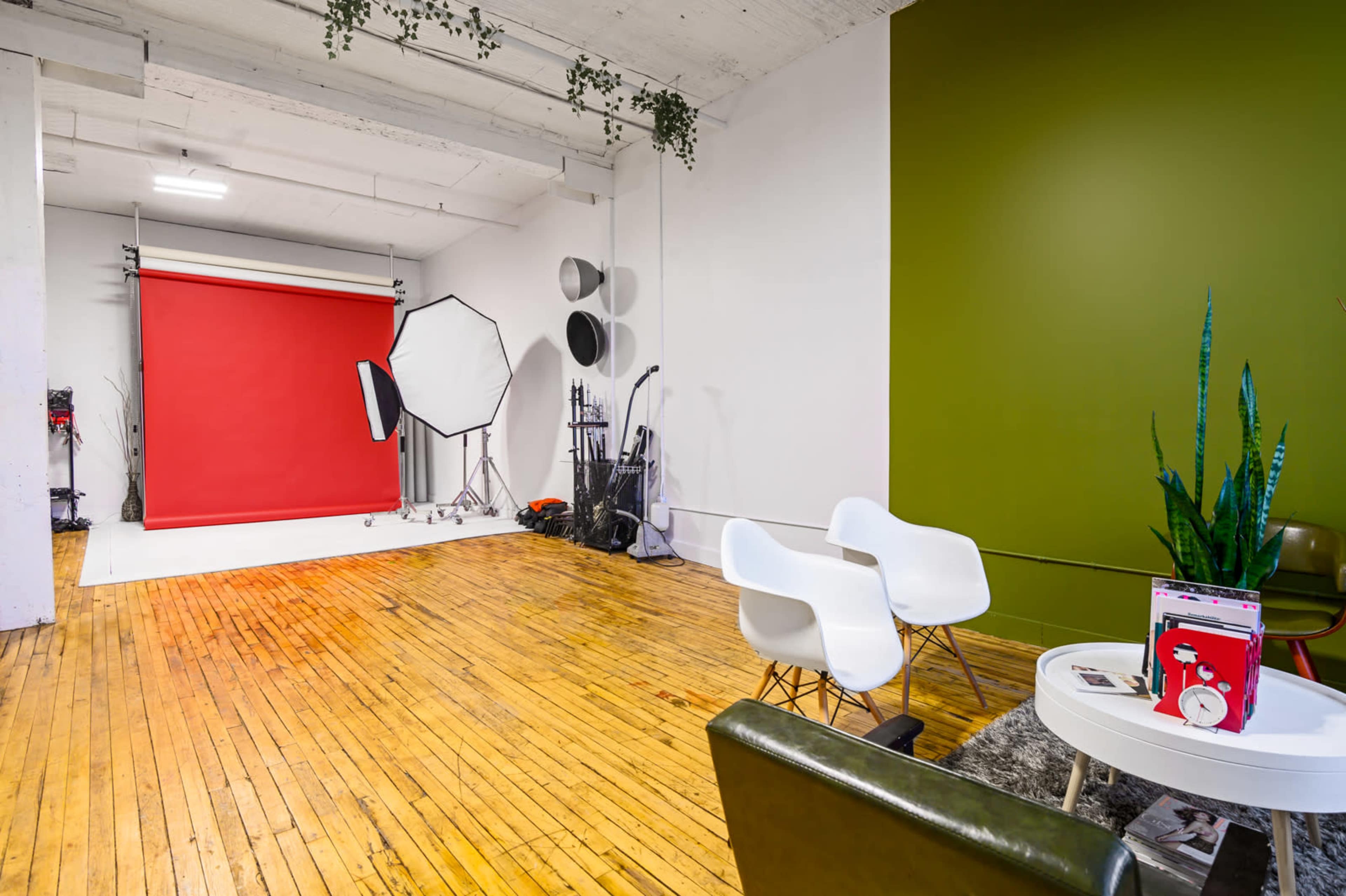 The image shows a photo studio with a red backdrop, softboxes, wooden floors, and a seating area featuring white chairs and a green wall.