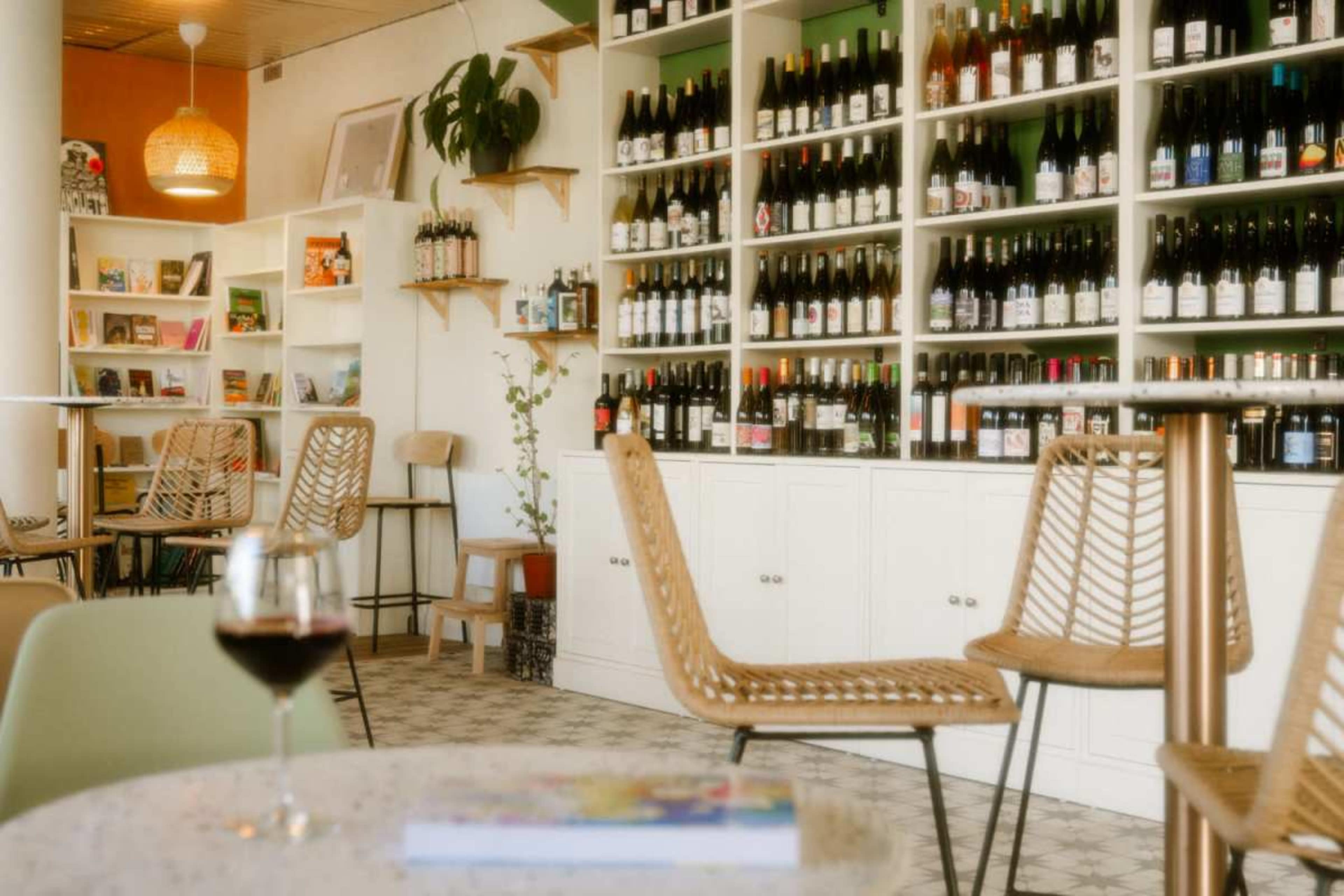 The image shows a cozy wine shop interior with shelves lined with wine bottles, wooden chairs, and a table with a glass of red wine.