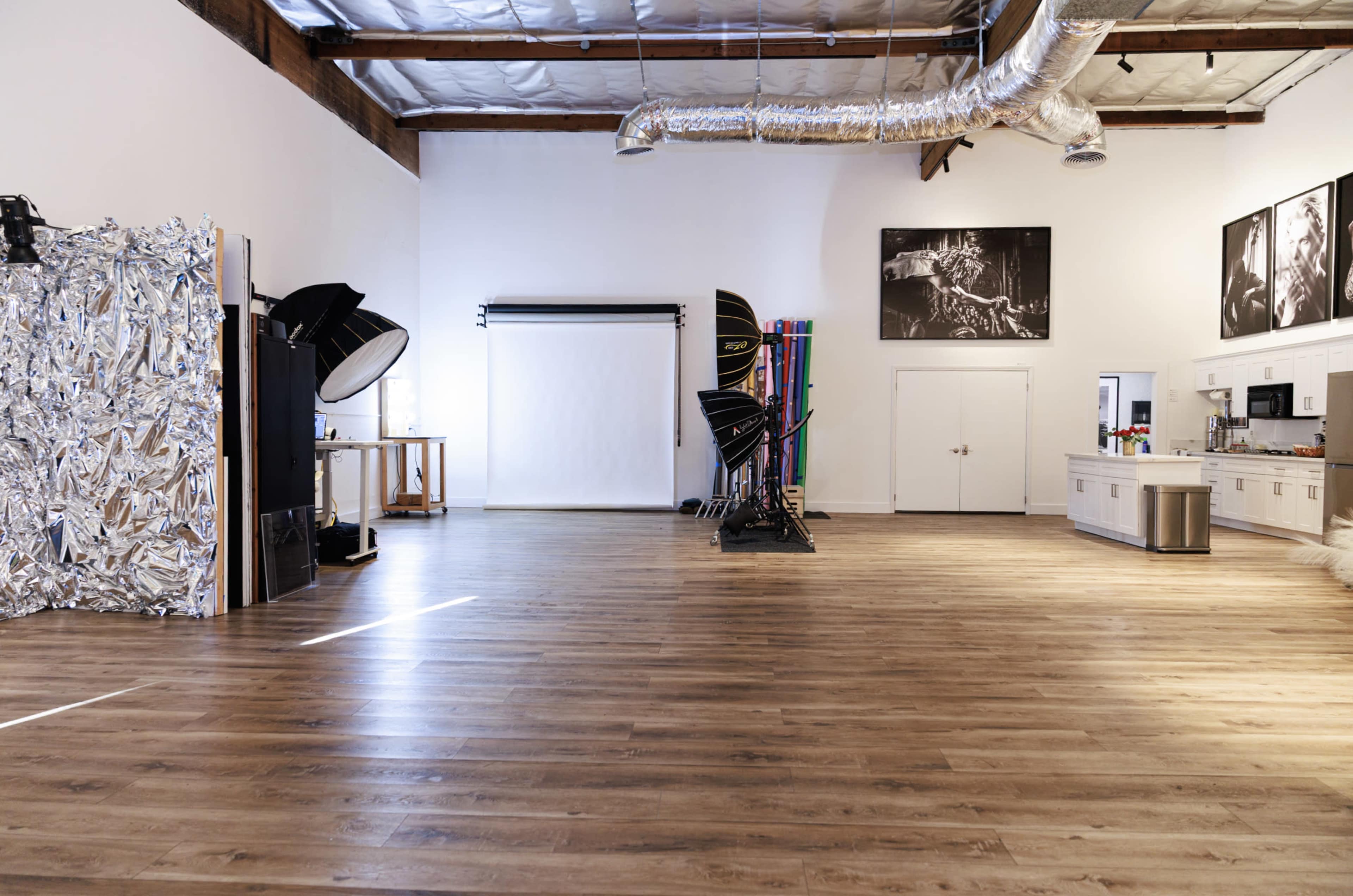 The image shows a spacious photography studio with wooden floors, a white backdrop, and various photography equipment positioned along one side.