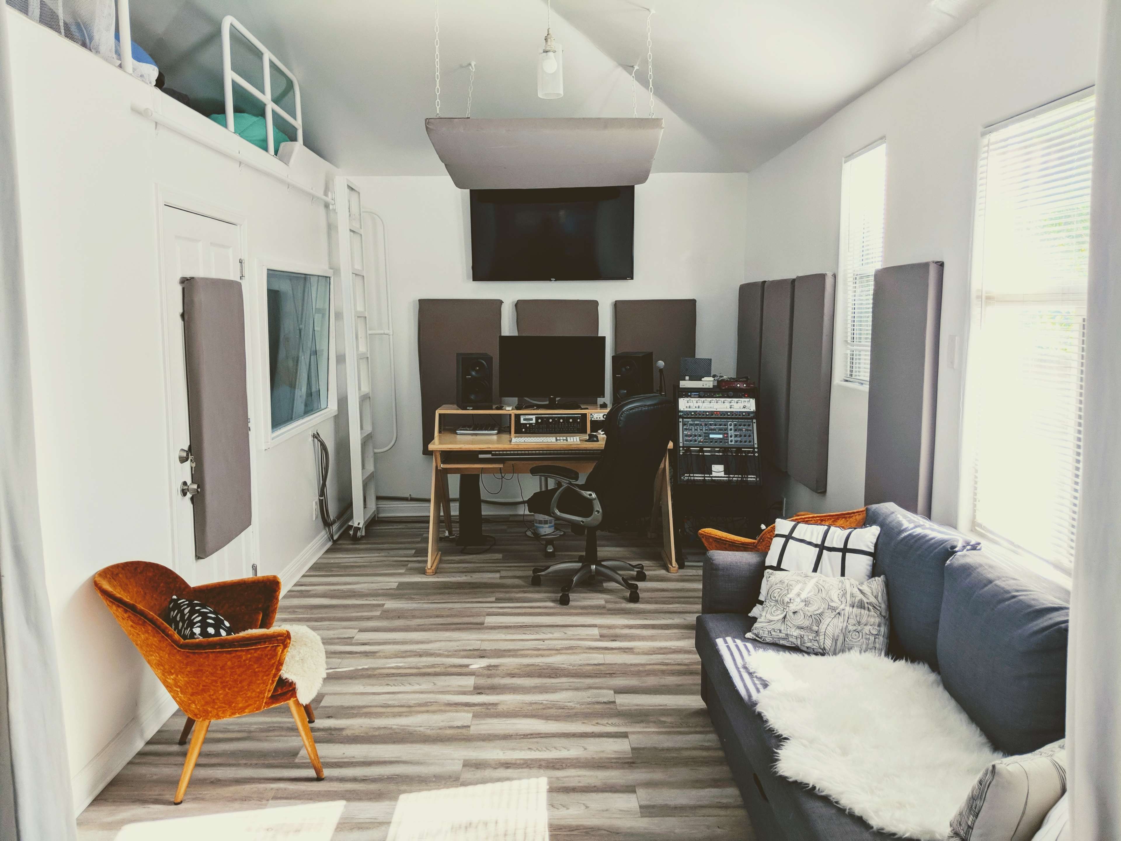 A cozy music studio featuring a desk with audio equipment, a comfortable couch, an armchair, and soundproof panels on the walls.