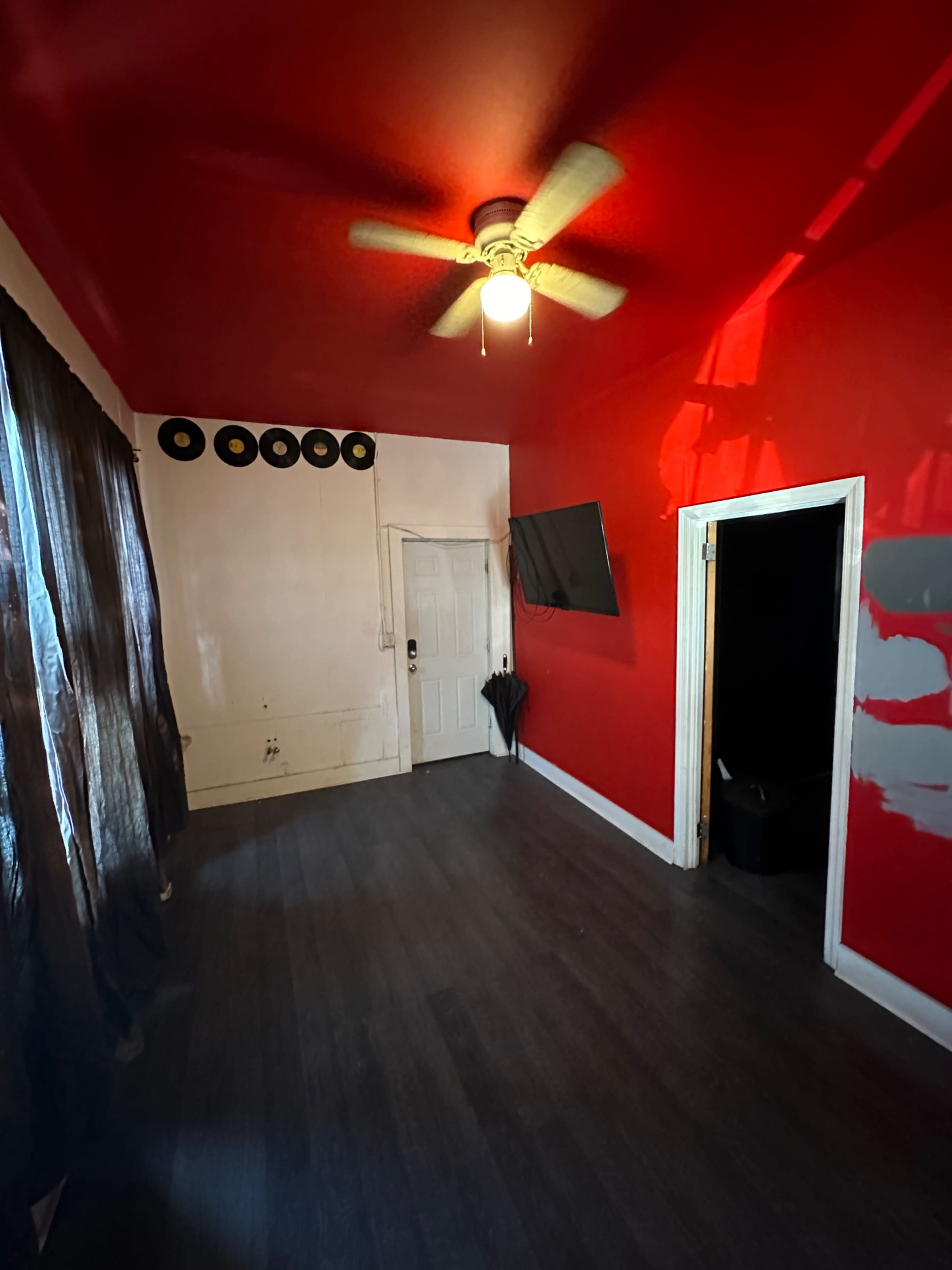The room features a red wall, a ceiling fan, dark curtains covering a window, a mounted TV, and a door leading to another space.