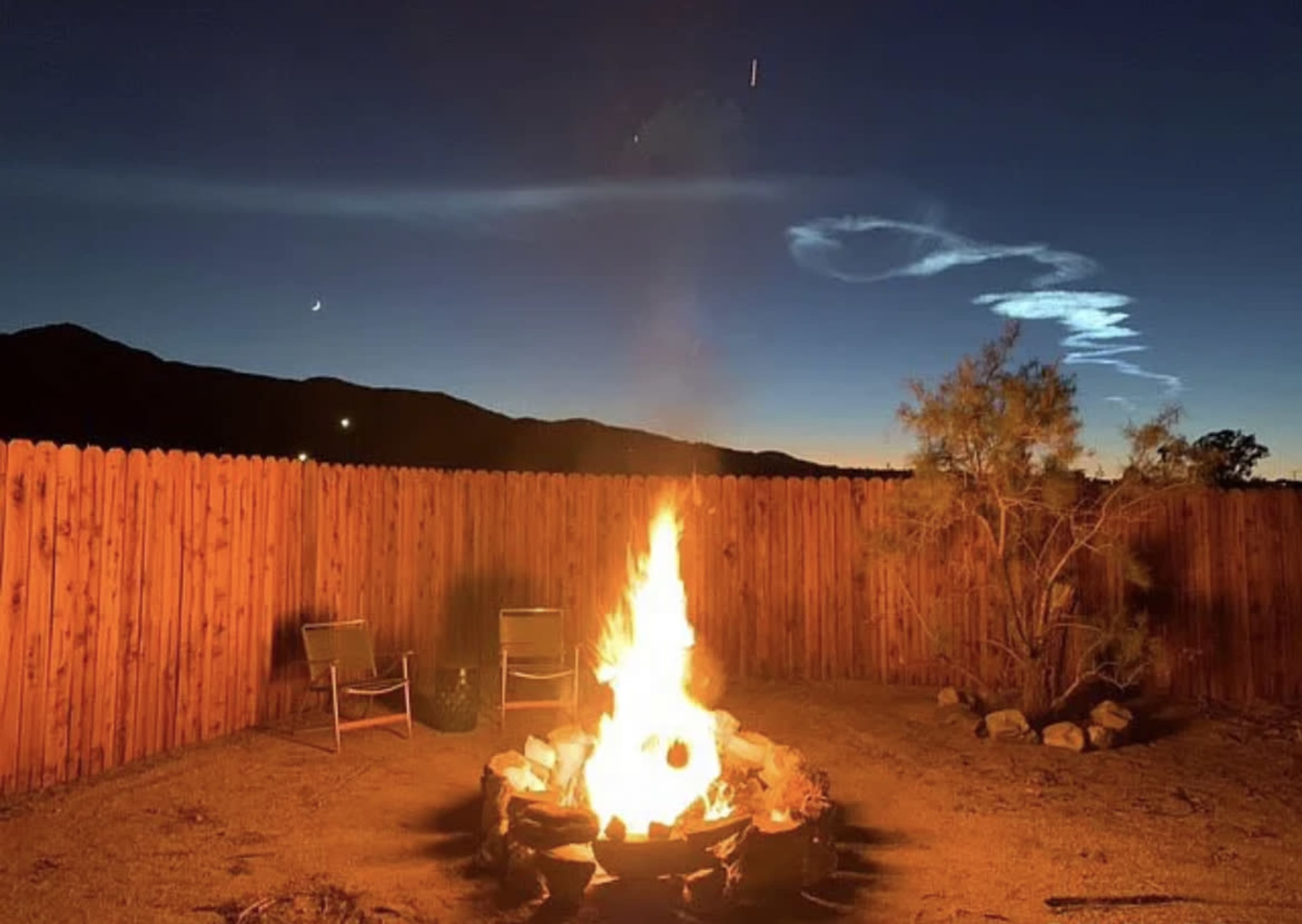 A campfire burns brightly in a desert backyard with a wooden fence, surrounded by chairs and silhouetted mountains against a twilight sky.