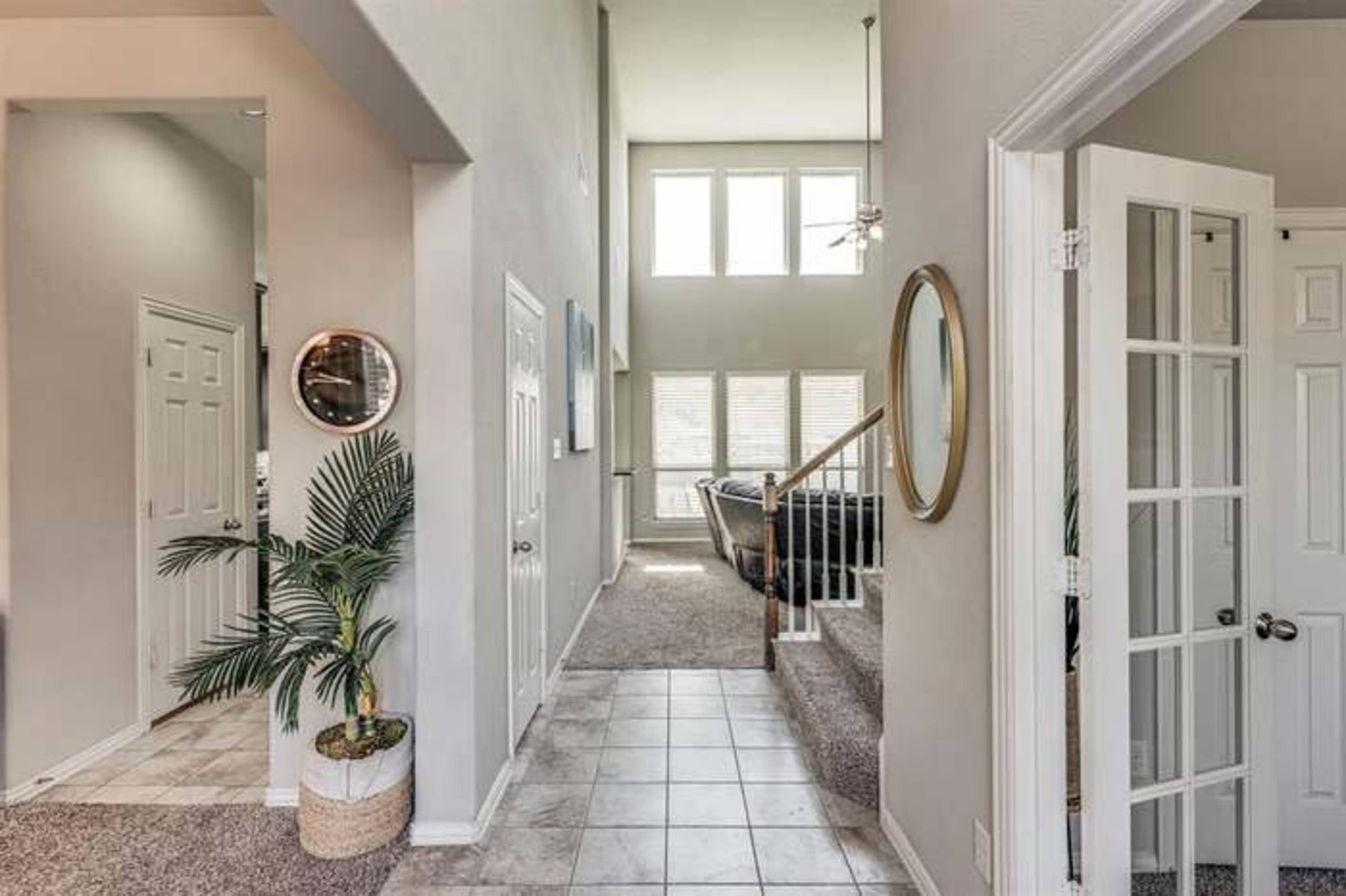 The image shows a bright entryway with a staircase leading to an upper level, decorated with a round mirror and a potted plant.