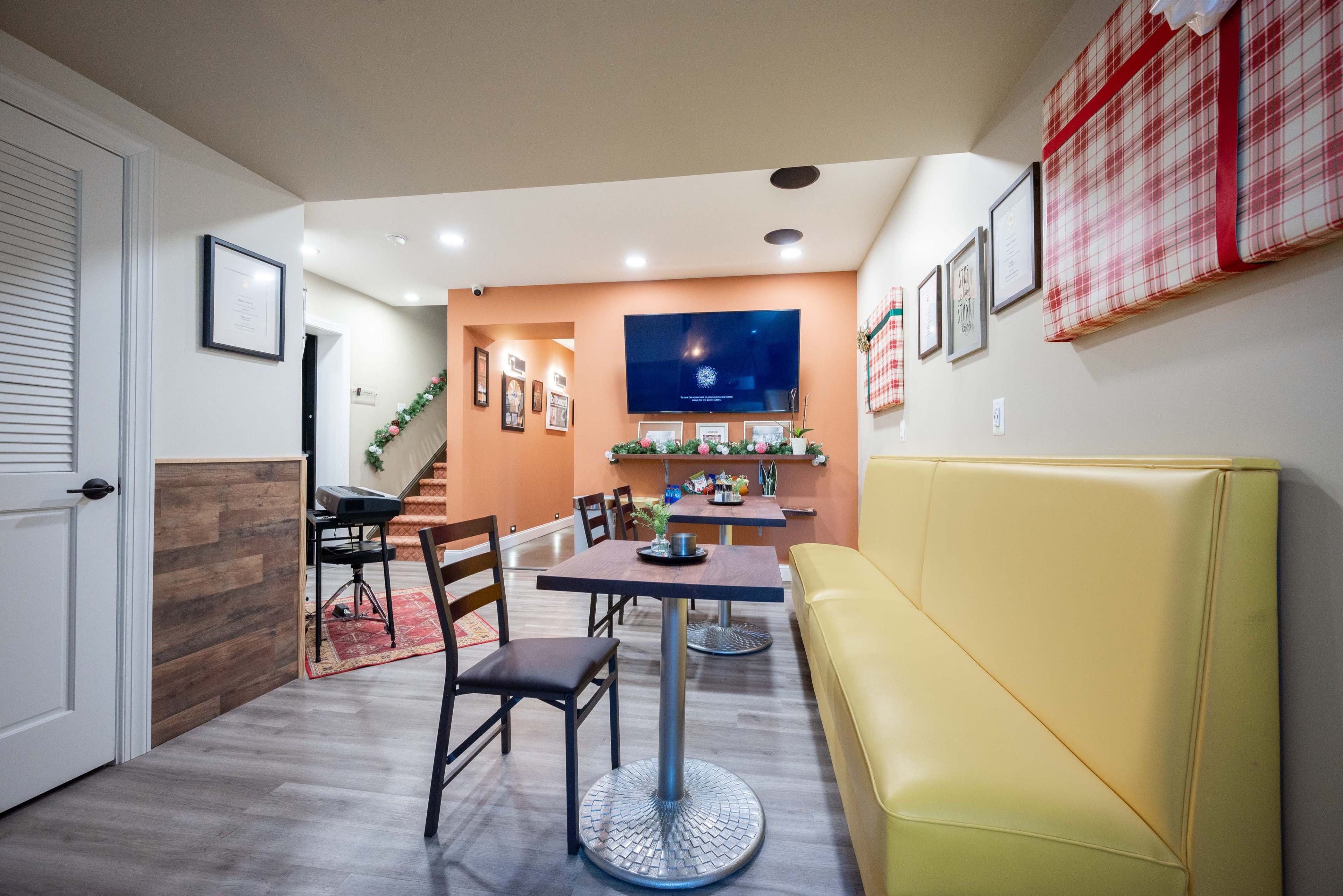 A cozy café interior featuring a yellow banquette, a small table with a chair, and a wall-mounted television, with stairs visible leading to another area.