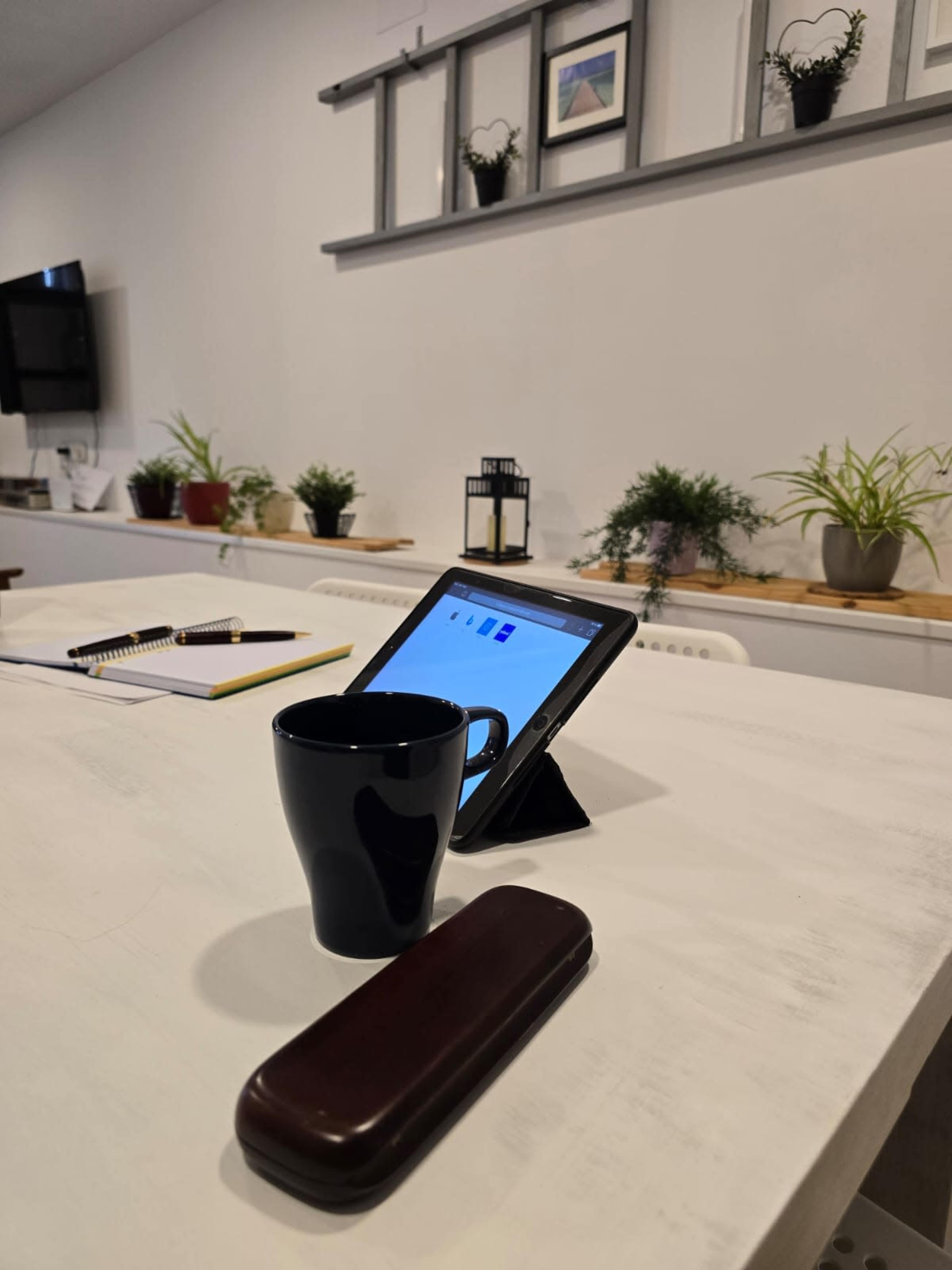 A tablet is positioned on a white table next to a black coffee cup and a wooden case, with a plant-filled background.
