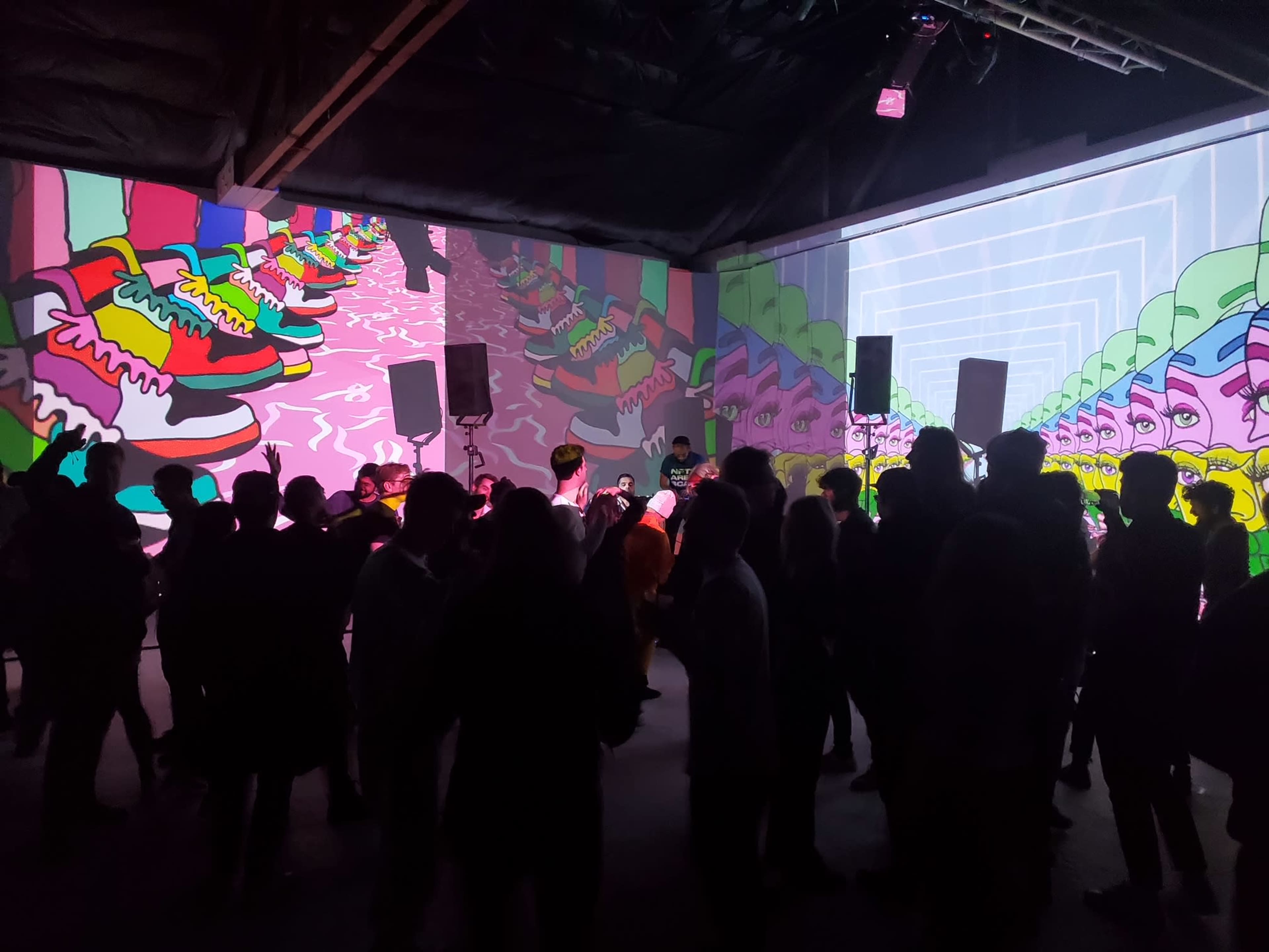 A crowd gathers in a dimly lit space featuring vibrant, animated projections of colorful shoes on the walls.