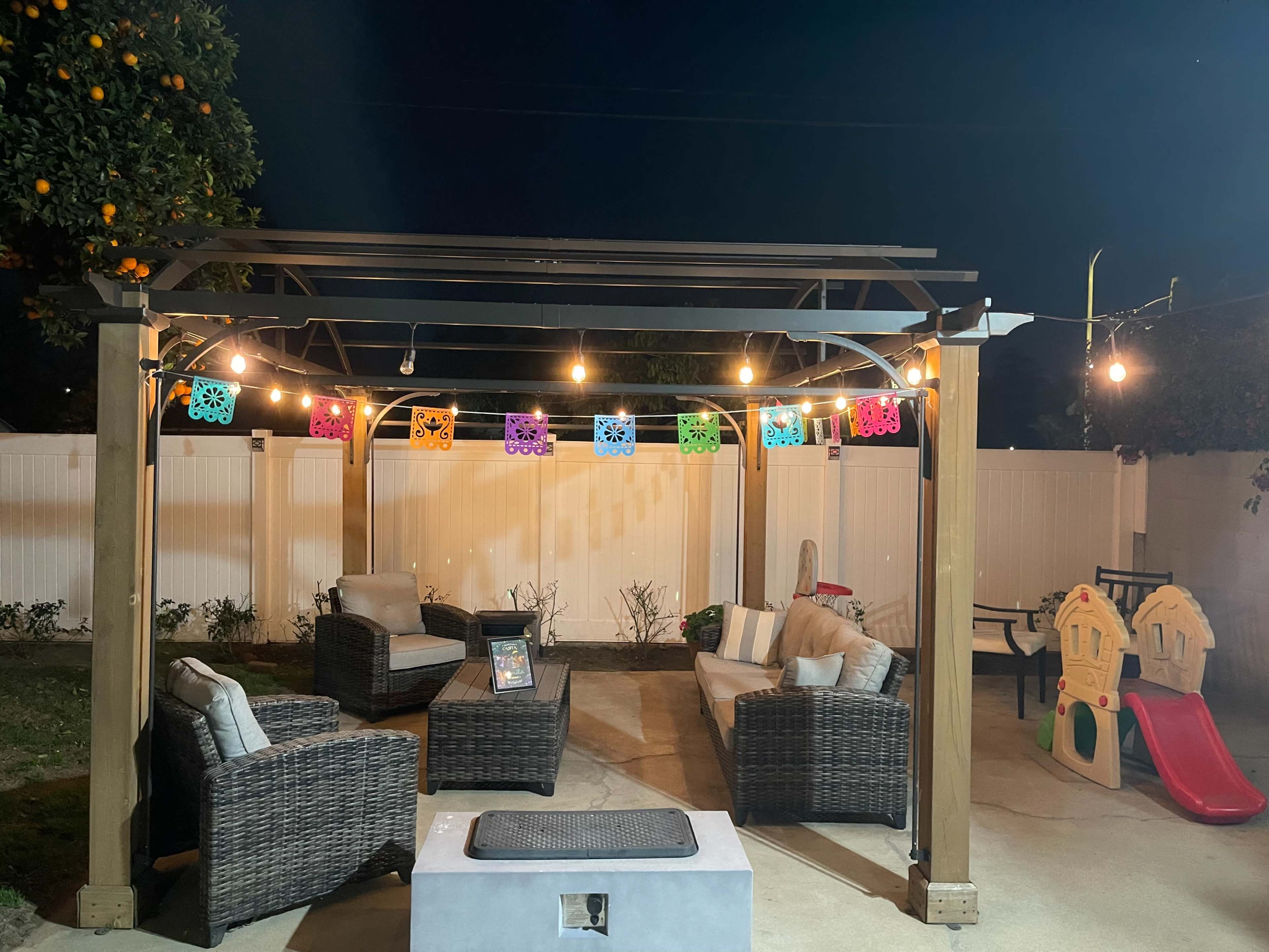 A patio with a pergola is illuminated by string lights and decorated with colorful papel picado banners, featuring seating arrangements and a children's play structure nearby.