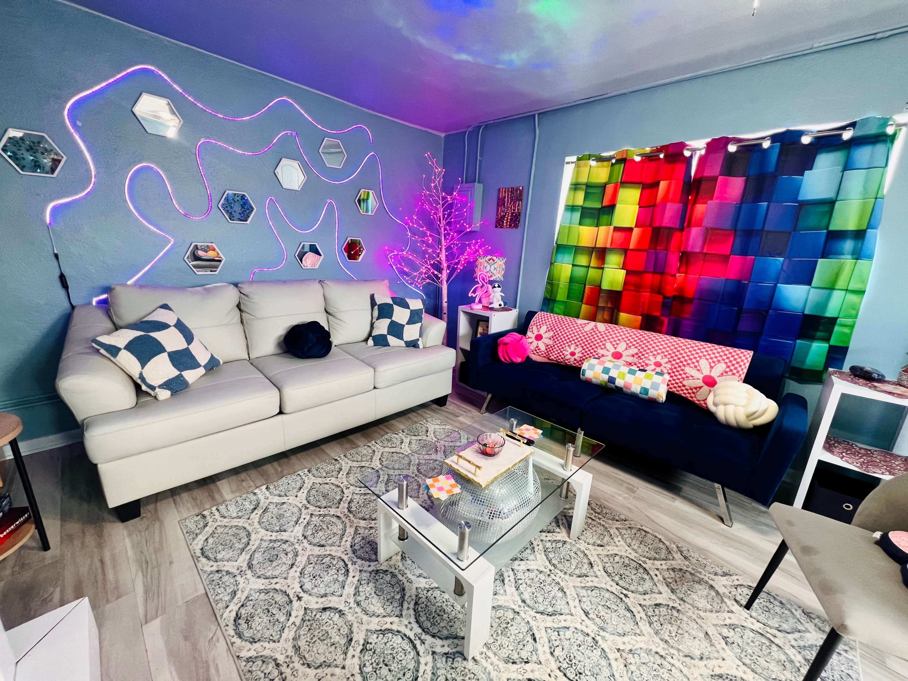 A colorful living room with a white sofa, a dark sofa adorned with cushions, a glass coffee table, and decorative wall art illuminated by colorful lights.