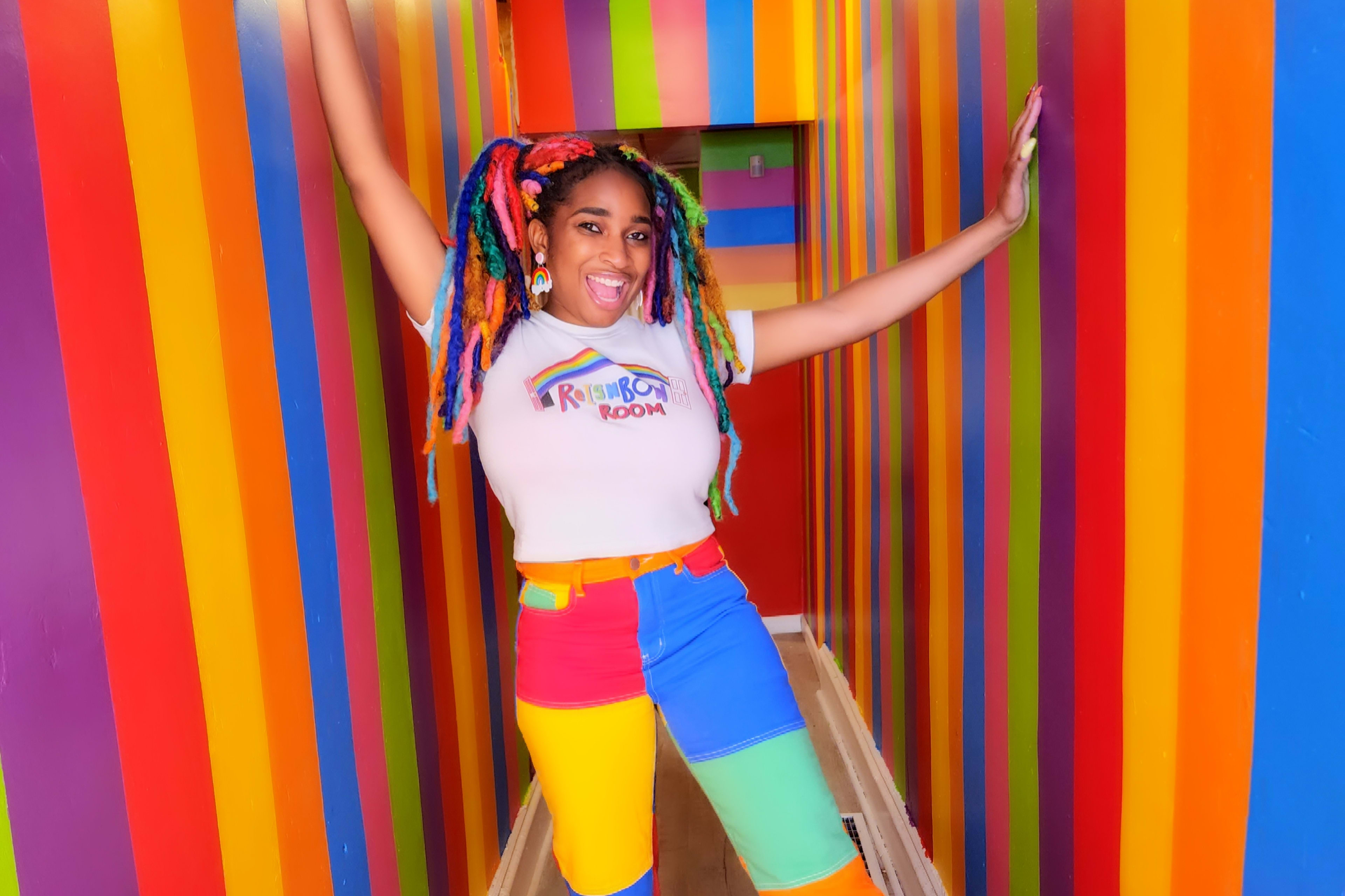A person with colorful, multi-colored hair poses in a brightly painted, striped hall.