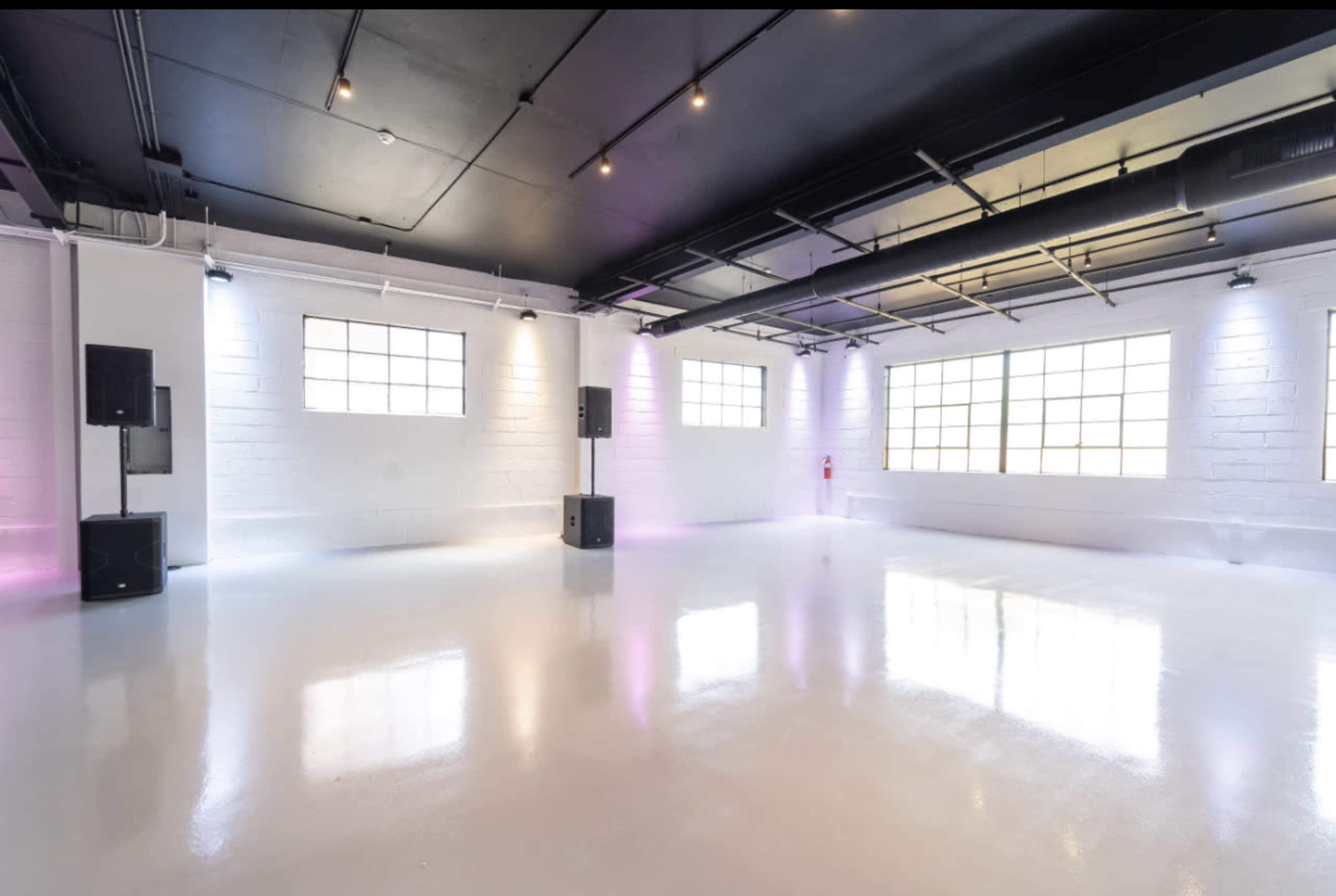 The image shows a spacious, empty room with polished white floors, large windows, and sound equipment positioned in the corners.