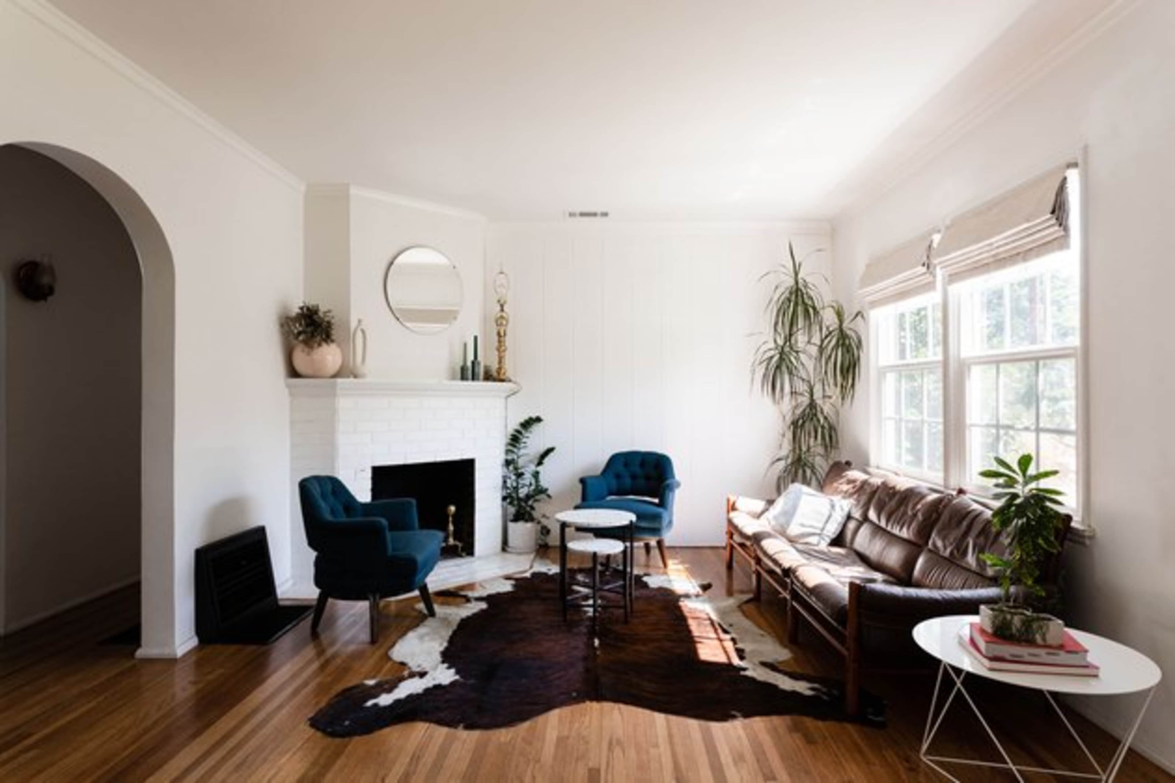A stylish living room features a brown leather sofa, two blue chairs, a round coffee table, and a white fireplace with a circular mirror above it.