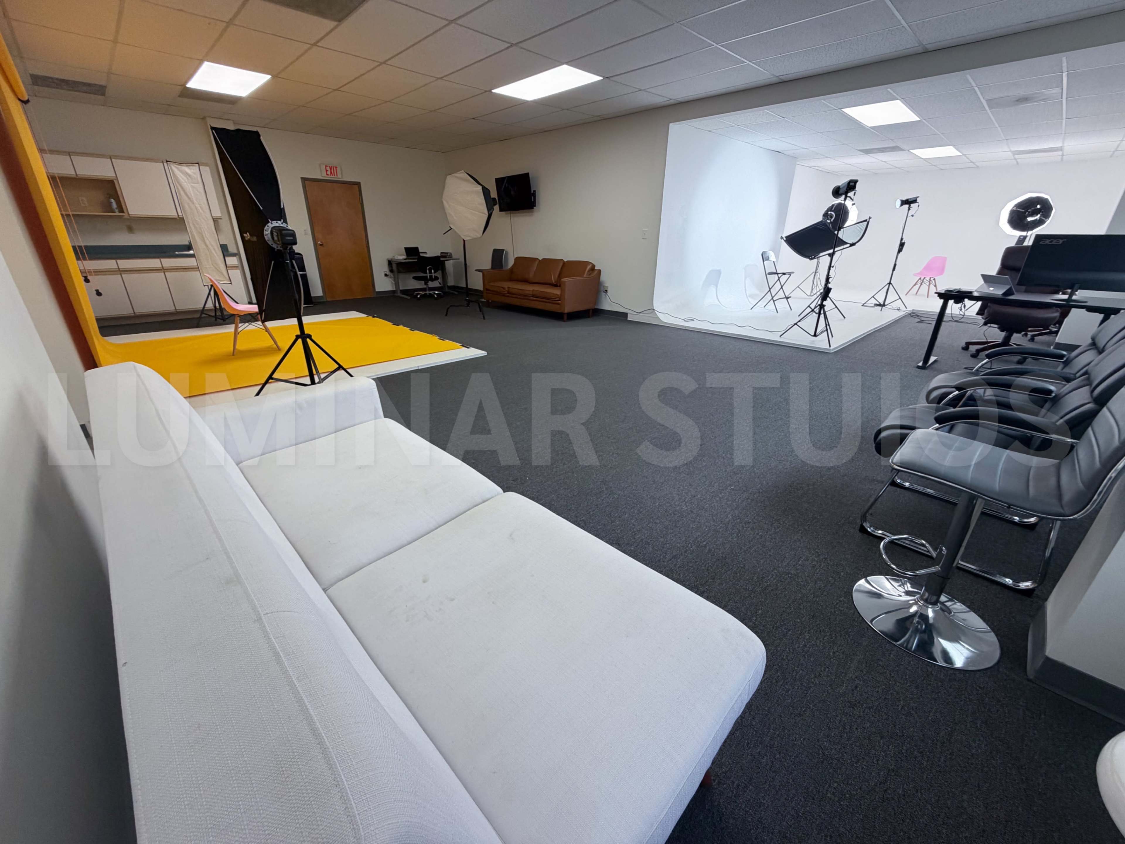 The image shows a spacious studio setup featuring a white backdrop, various photography equipment, a couch, and seating arrangements.
