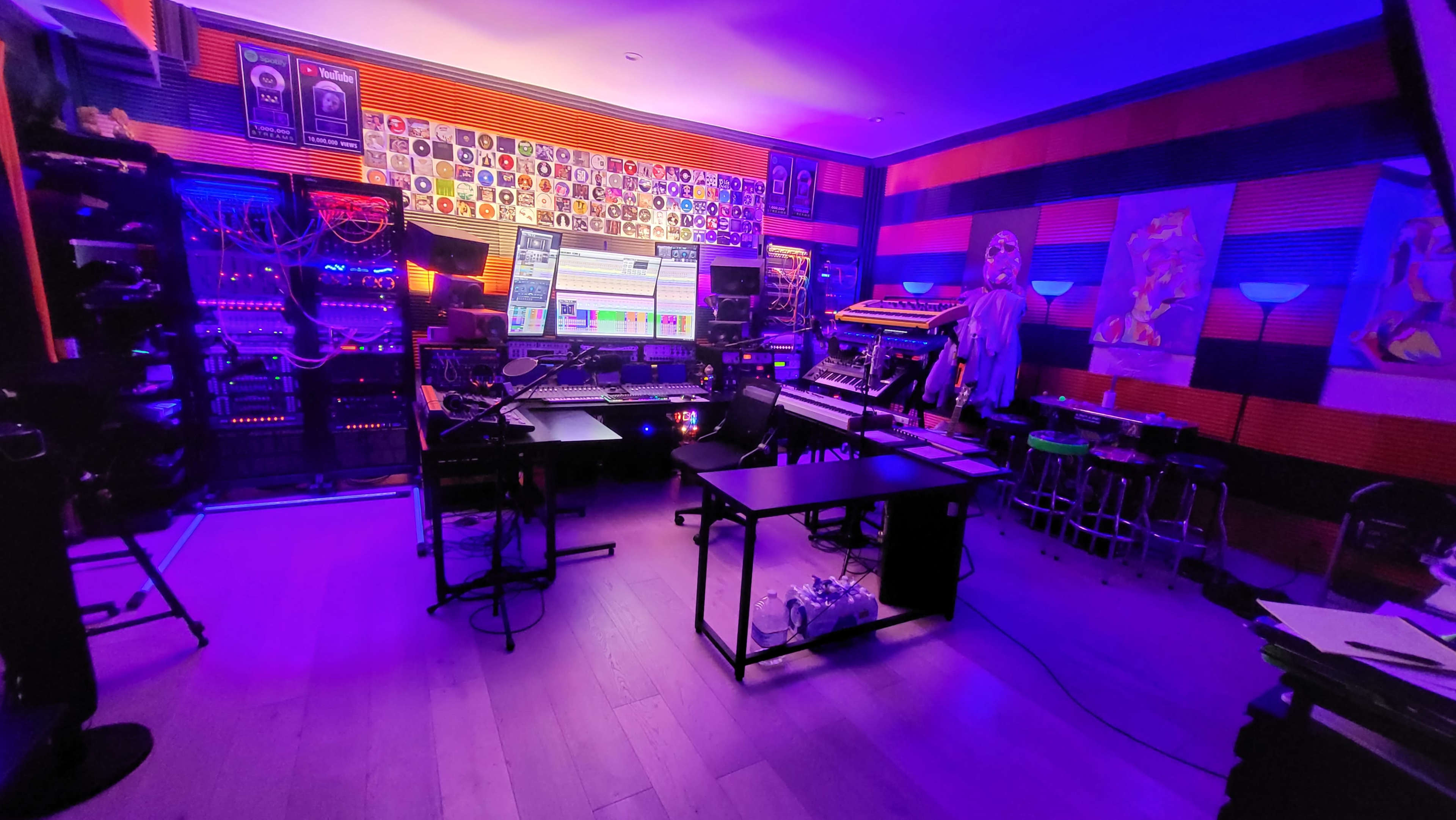 The image shows a music studio with colorful lighting, featuring various audio equipment, a large screen displaying a music production software, and decorated walls with artwork and vinyl records.
