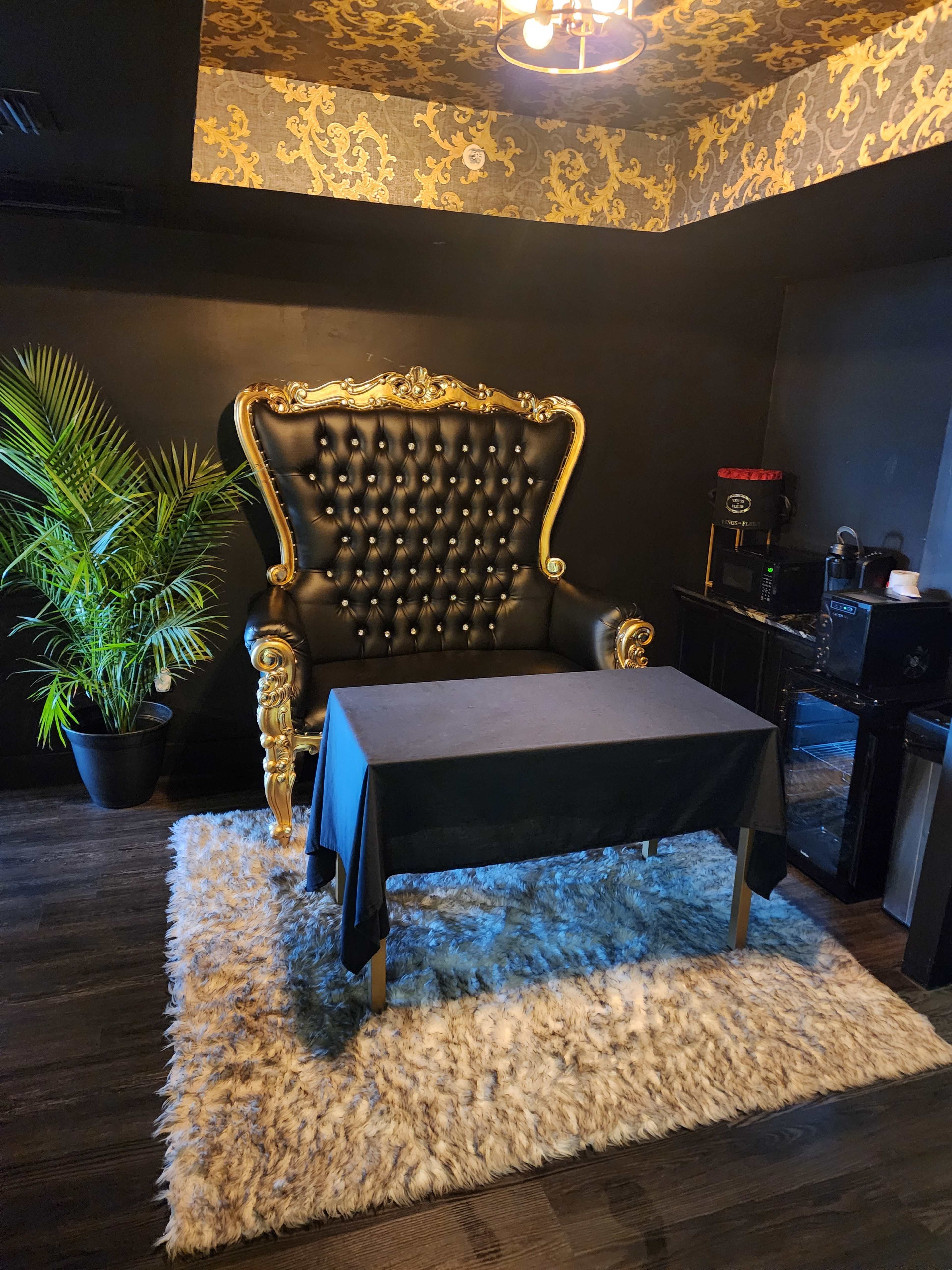 The image features a large, ornate black and gold throne chair next to a small table covered with a dark cloth, set on a plush rug in a dimly lit room with decorative wallpaper.