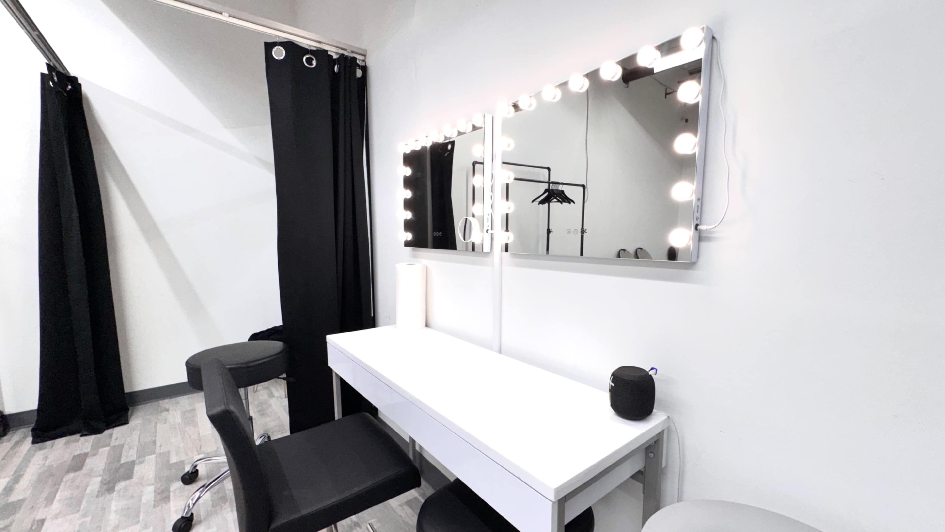 A brightly lit vanity mirror is positioned above a white table in a room with black curtains and chairs.