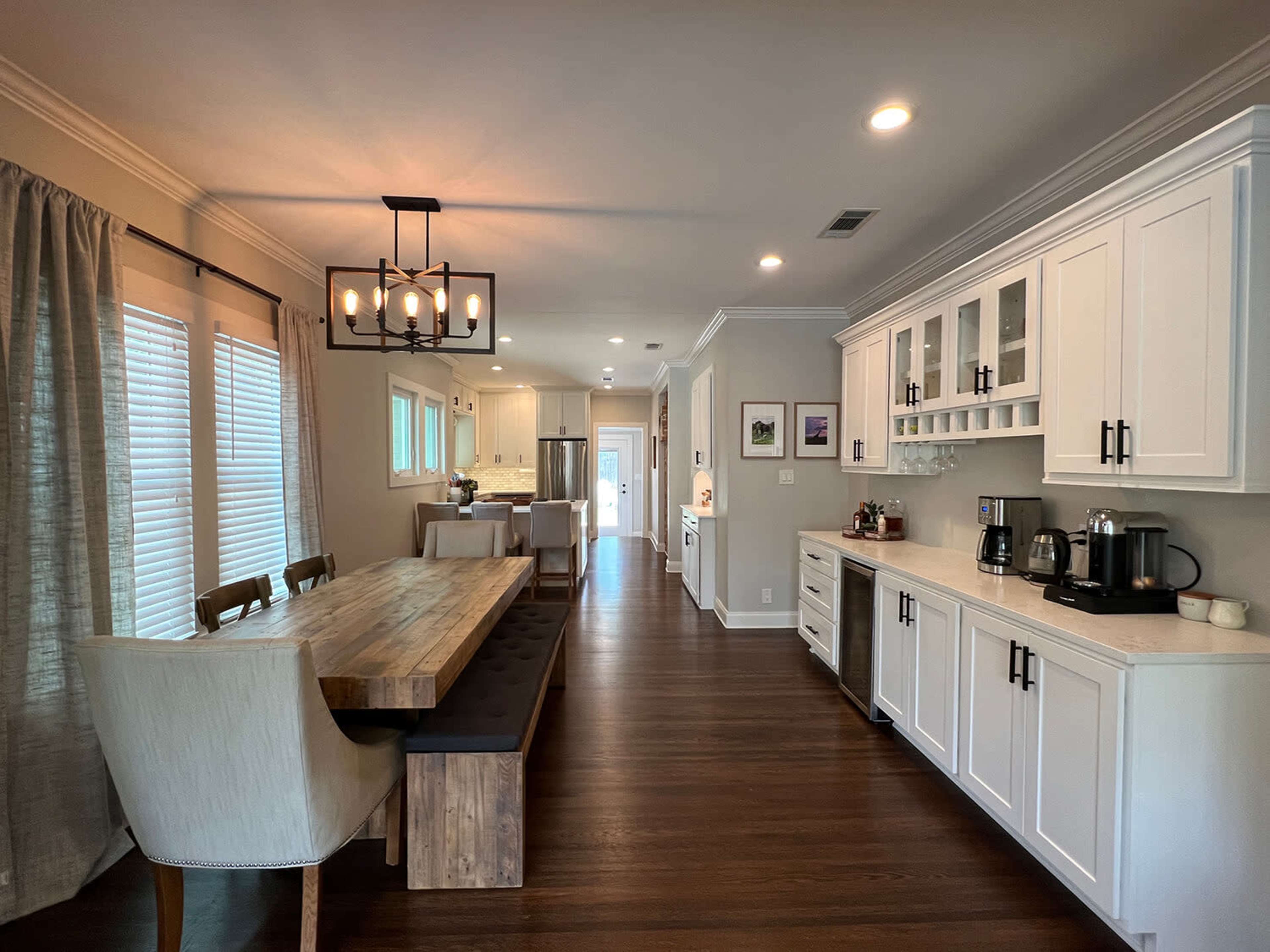 A modern kitchen features a long wooden table with bench seating, complemented by white cabinetry and stainless steel appliances.