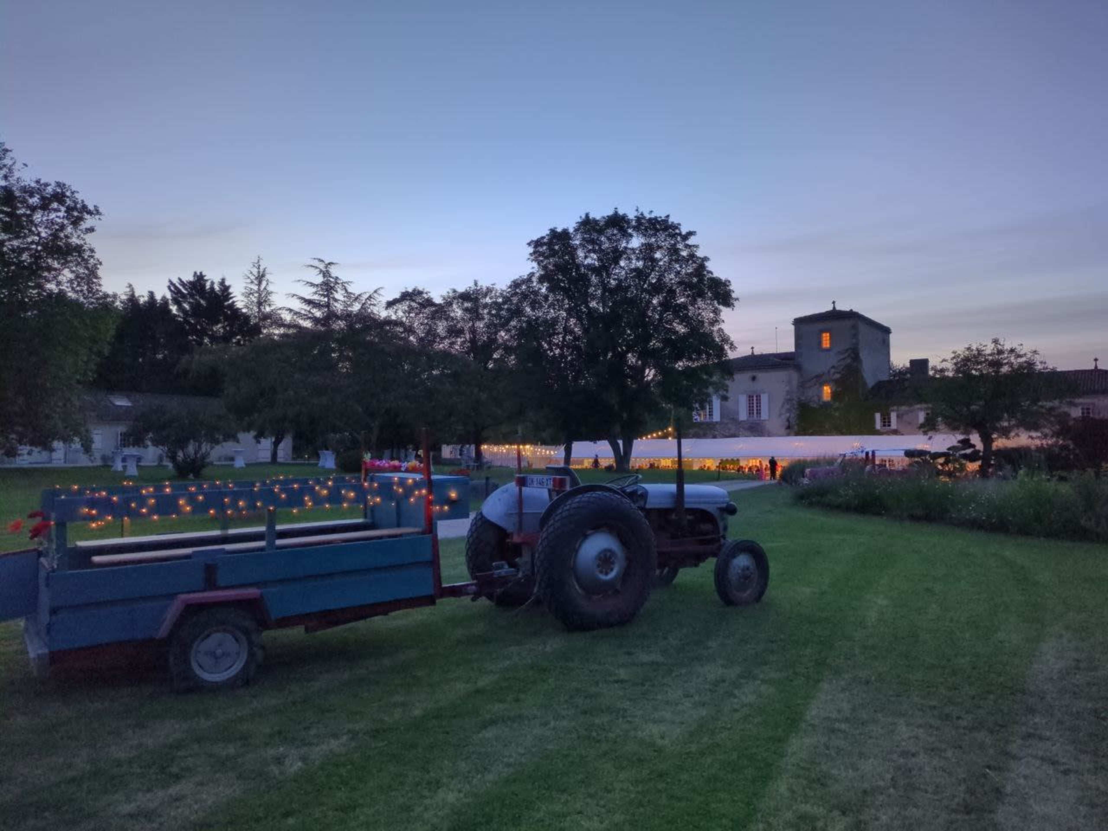 A tractor with a trailer is parked on a grassy field near a building adorned with string lights, set against a twilight sky.