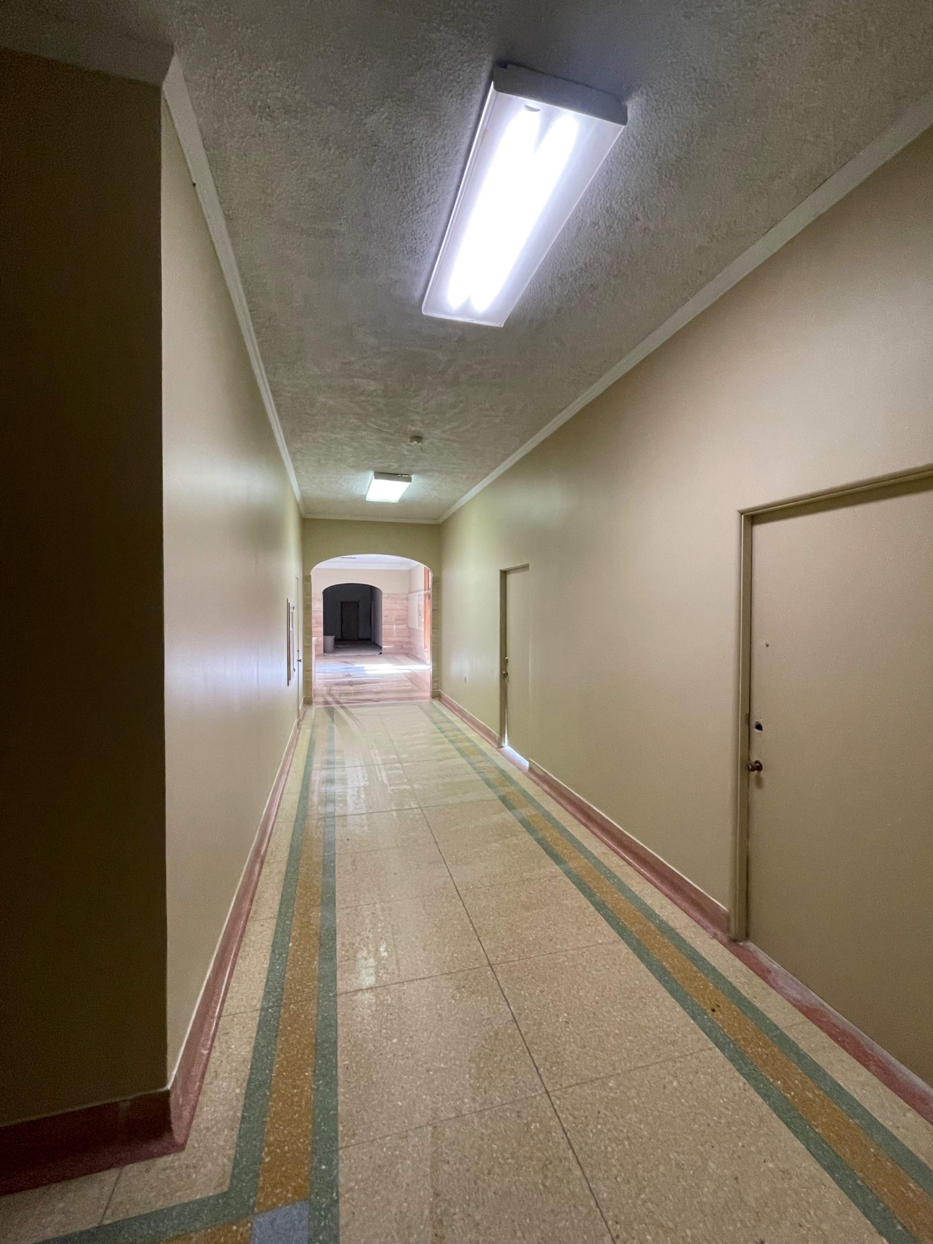 A long, narrow corridor with beige walls and a tiled floor leads to an archway at the end.