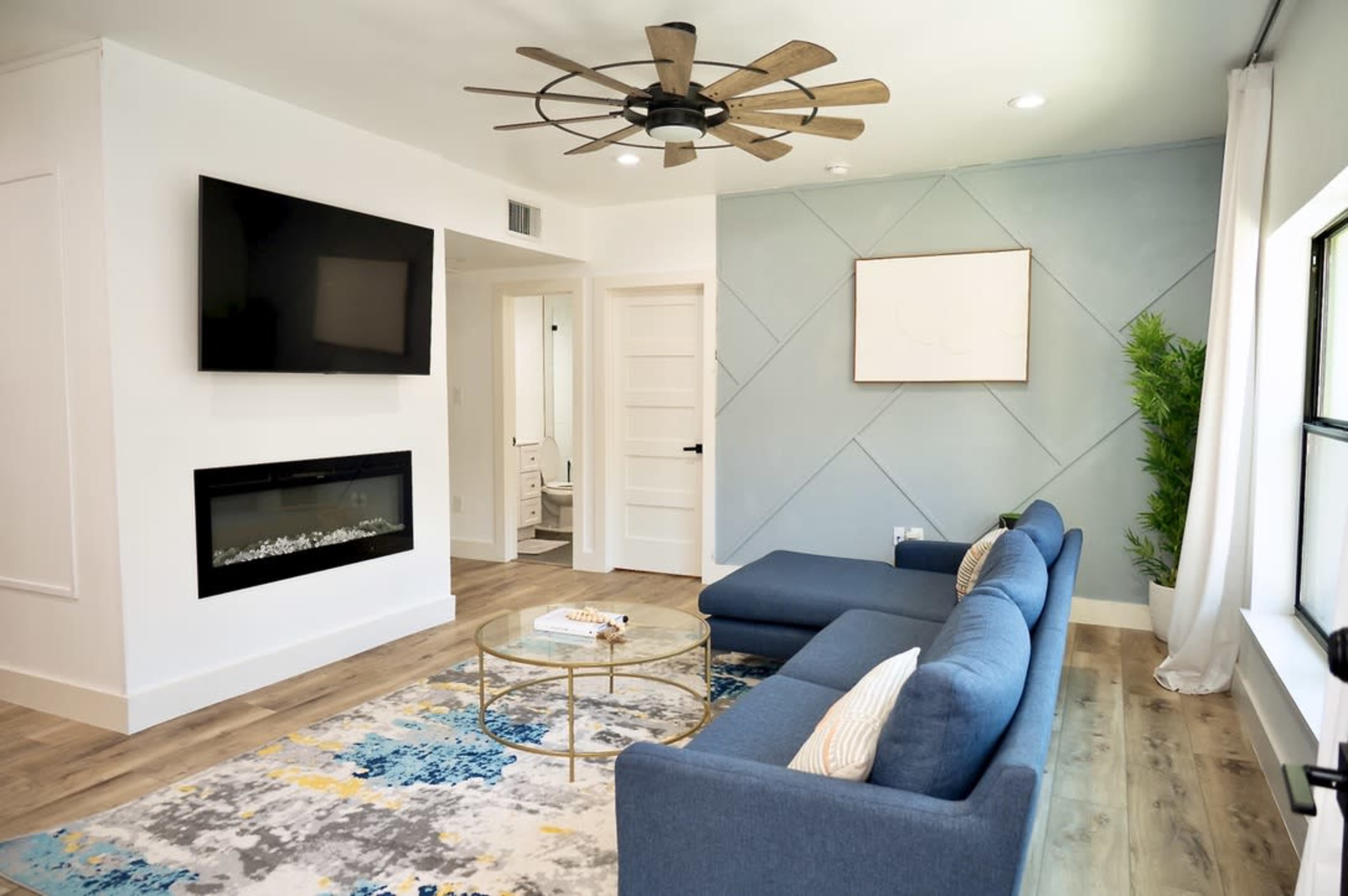 The image shows a modern living room with a blue sectional sofa, a wall-mounted television, an electric fireplace, and a coffee table on a patterned rug.