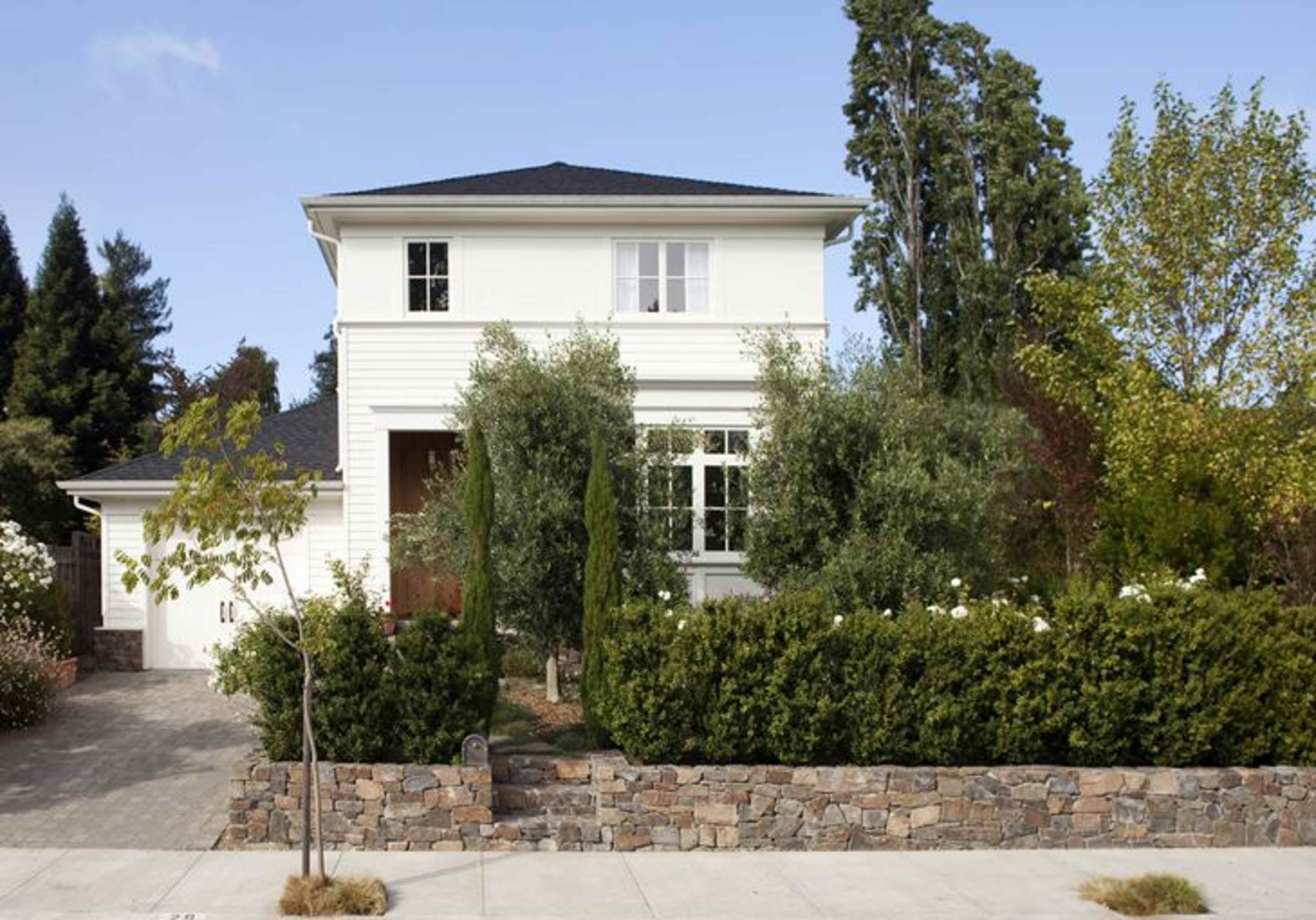 A two-story white house surrounded by trees and shrubs, with a stone wall in the front.