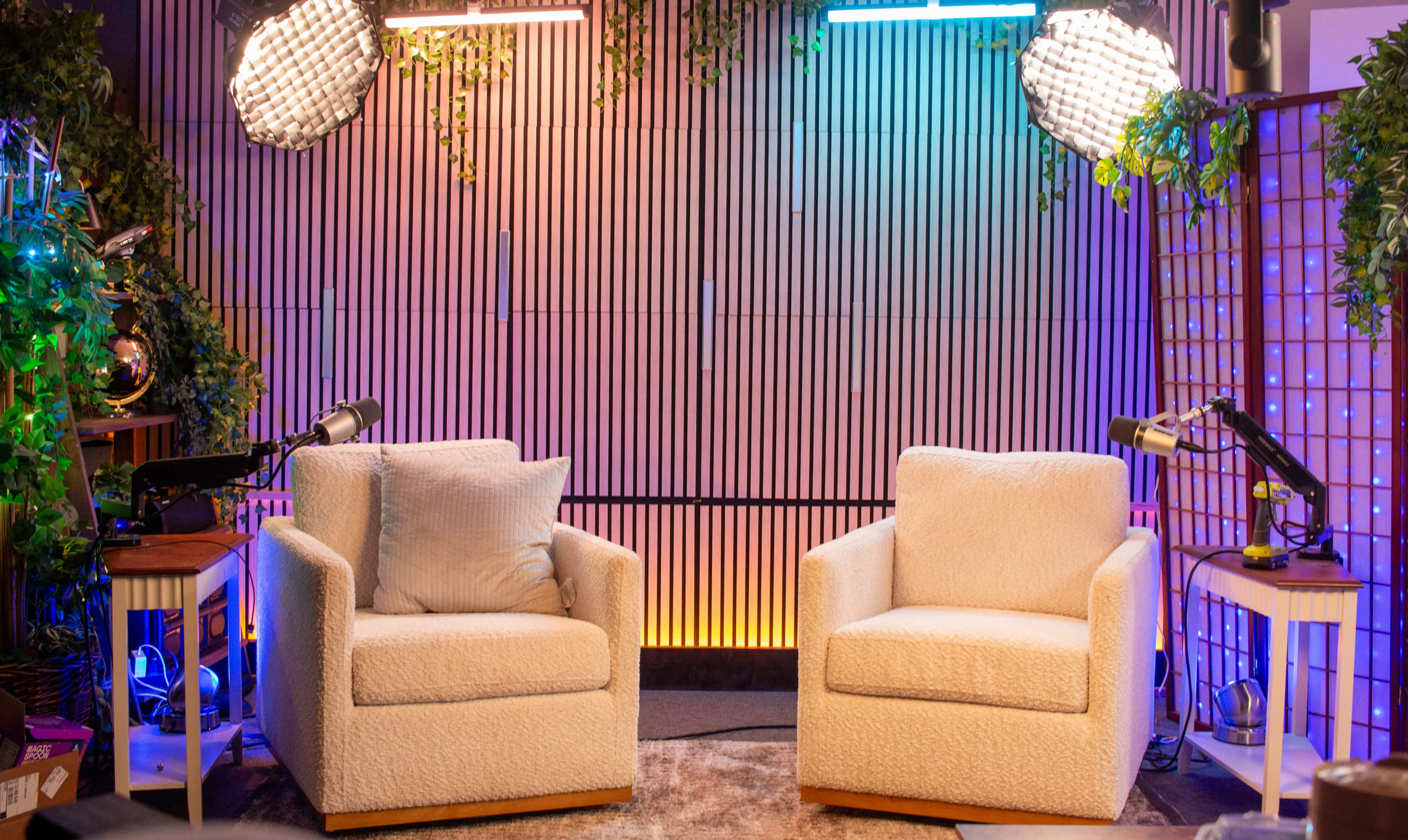 The image shows a brightly lit studio setup featuring two white upholstered chairs positioned facing each other against a backdrop of wooden slats with colorful lighting.