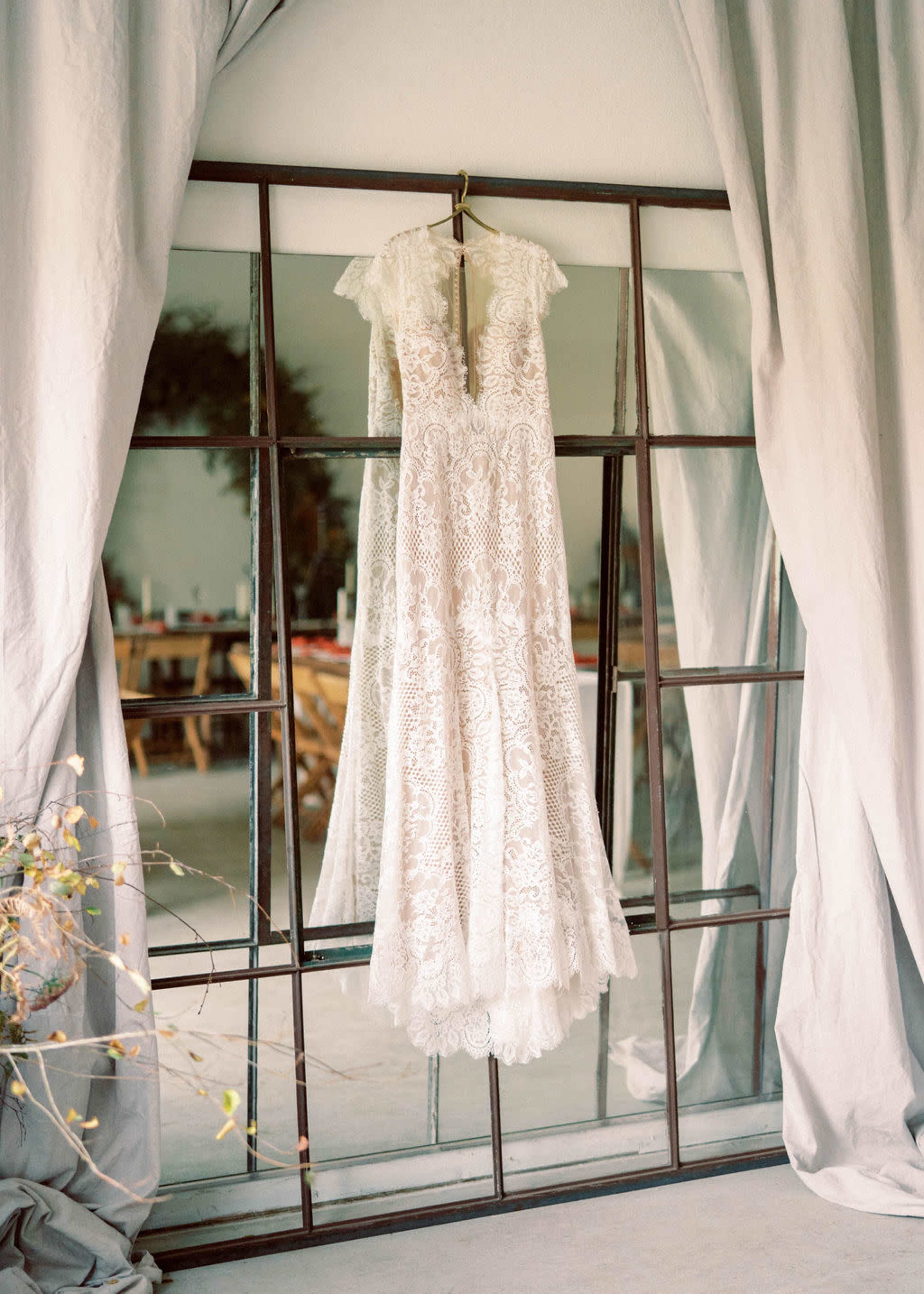 A lace wedding dress hangs from a hook in front of a mirror, framed by a large window structure.