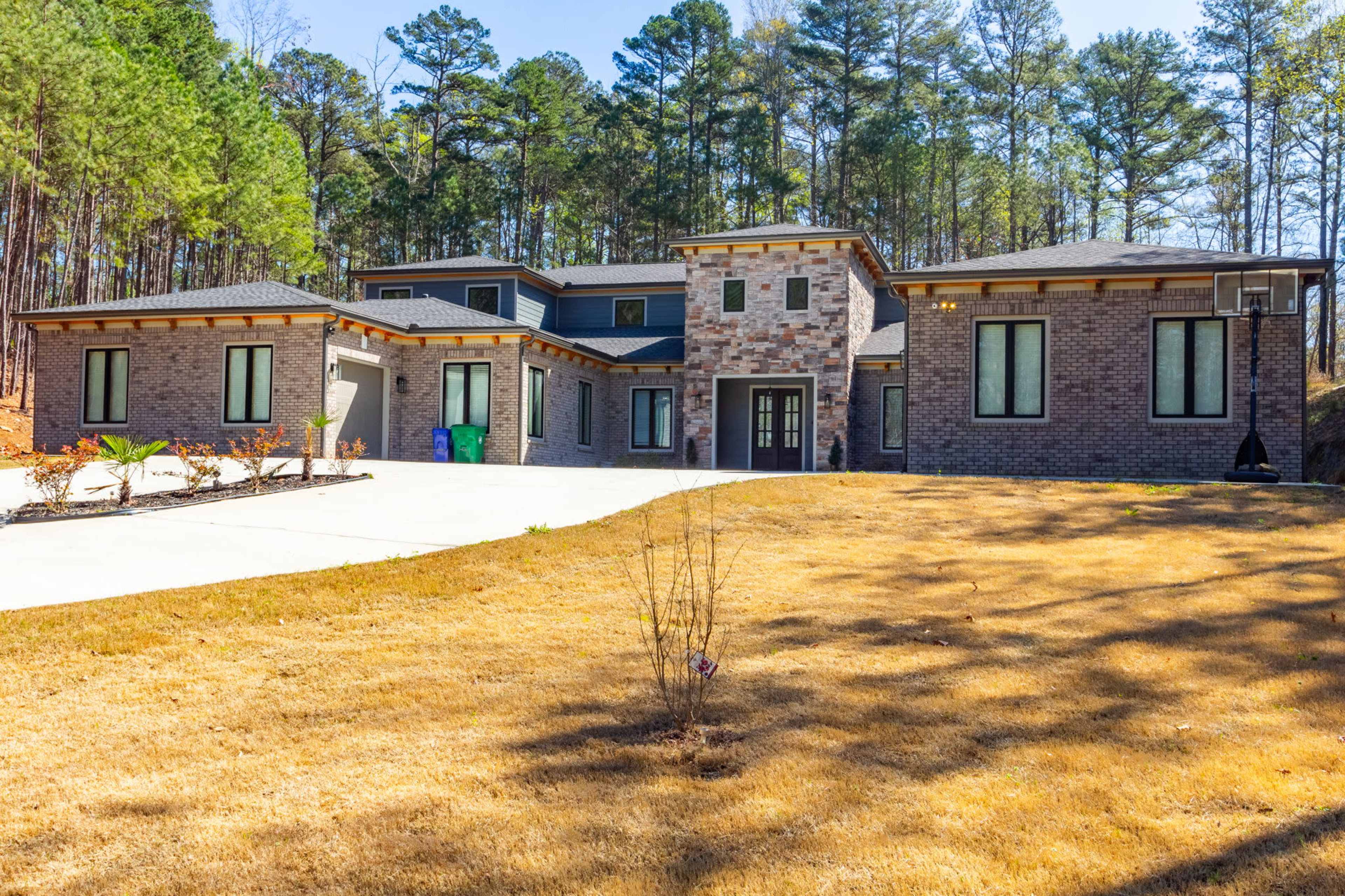 A modern two-story house with a brick exterior and large windows is situated at the end of a driveway, surrounded by pine trees.