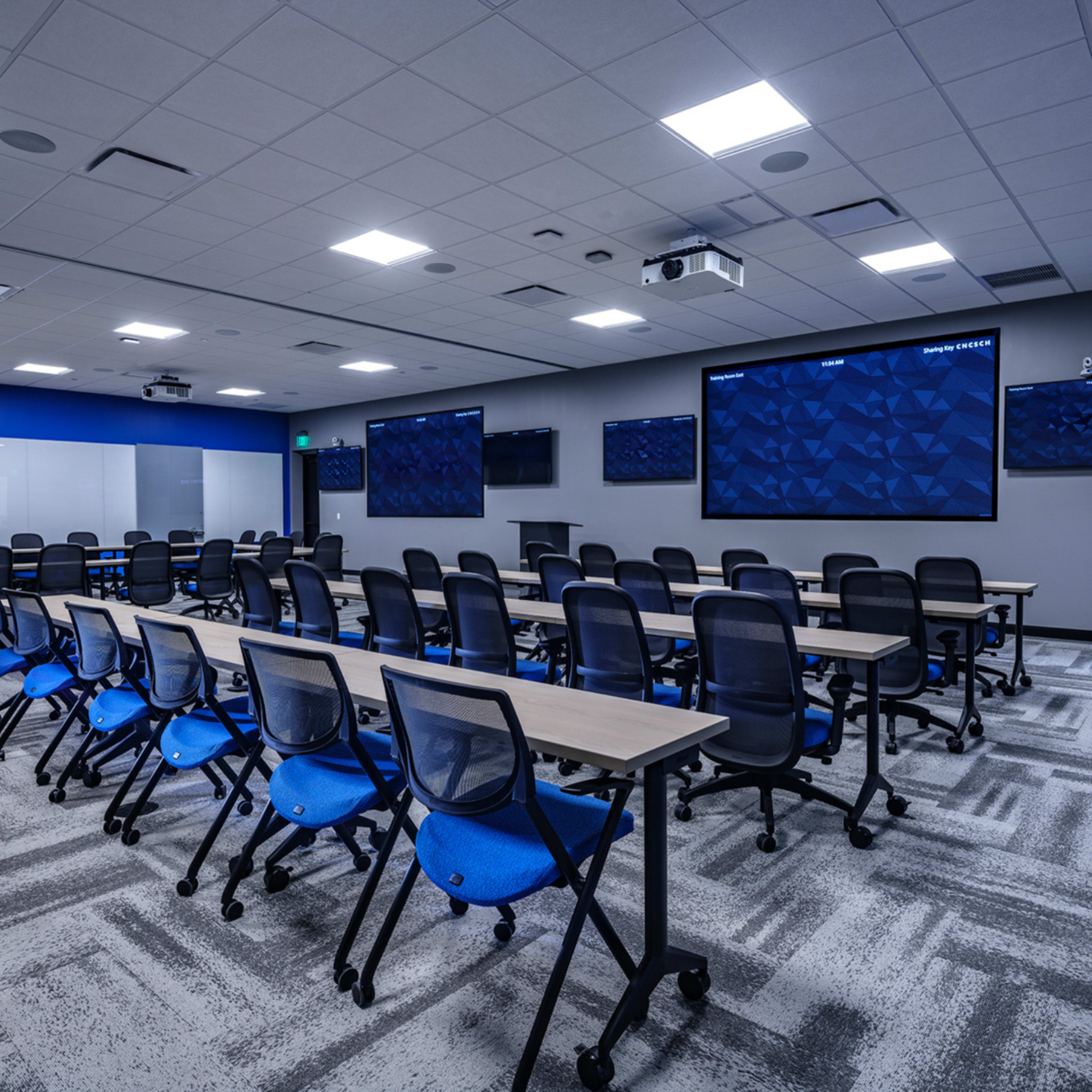 The image shows a modern classroom equipped with multiple rows of tables and chairs, large screens on the walls, and various high-tech installations.