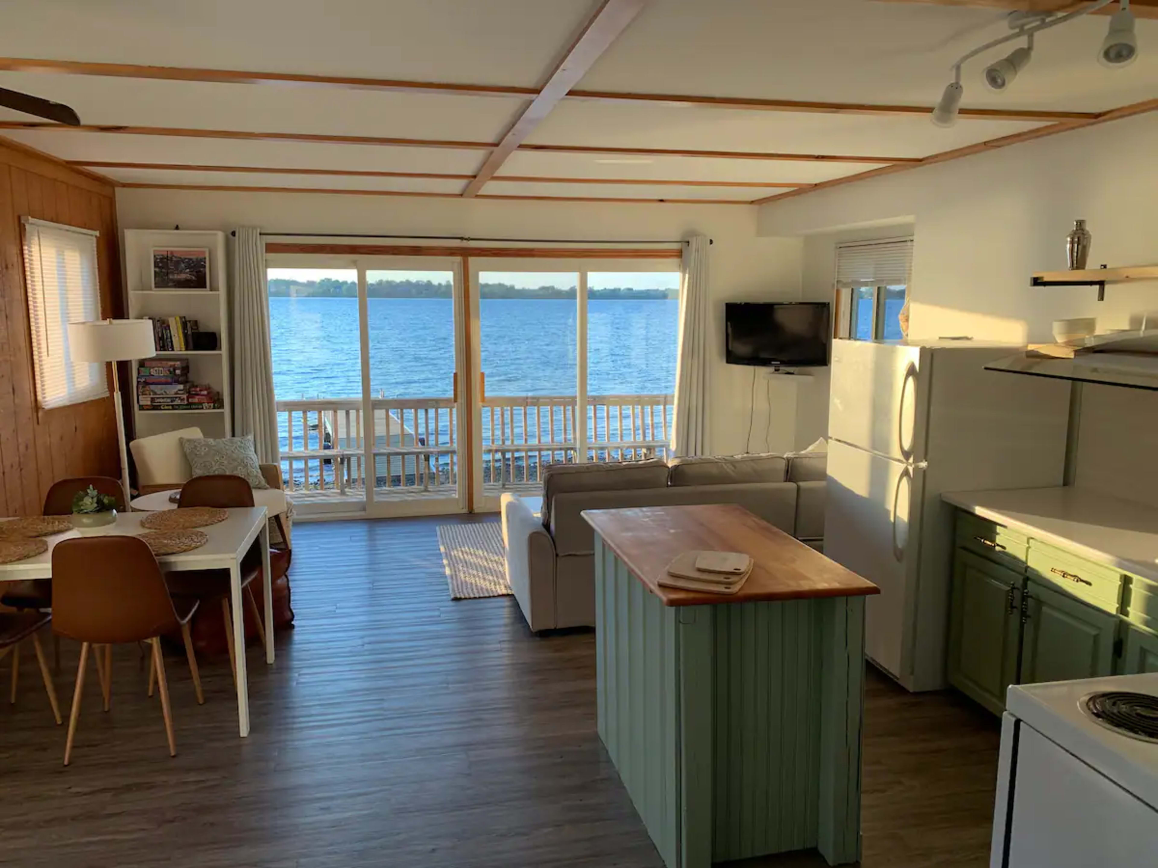 The image depicts a bright, modern living area with a kitchen island, dining table, and large windows overlooking a body of water.