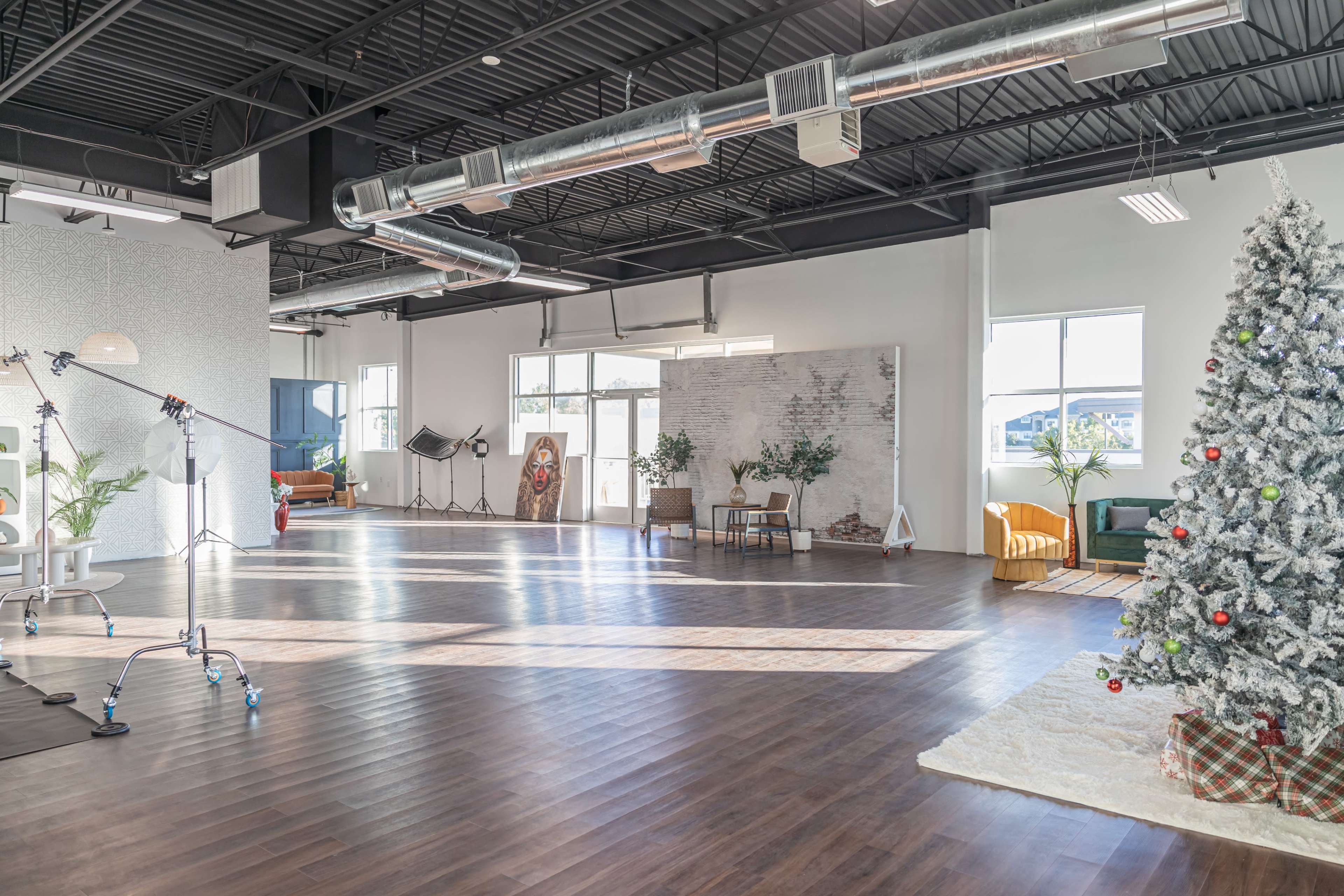 The image shows a spacious, well-lit studio with wooden flooring, large windows, a decorated Christmas tree, and various furniture and greenery arranged throughout the space.