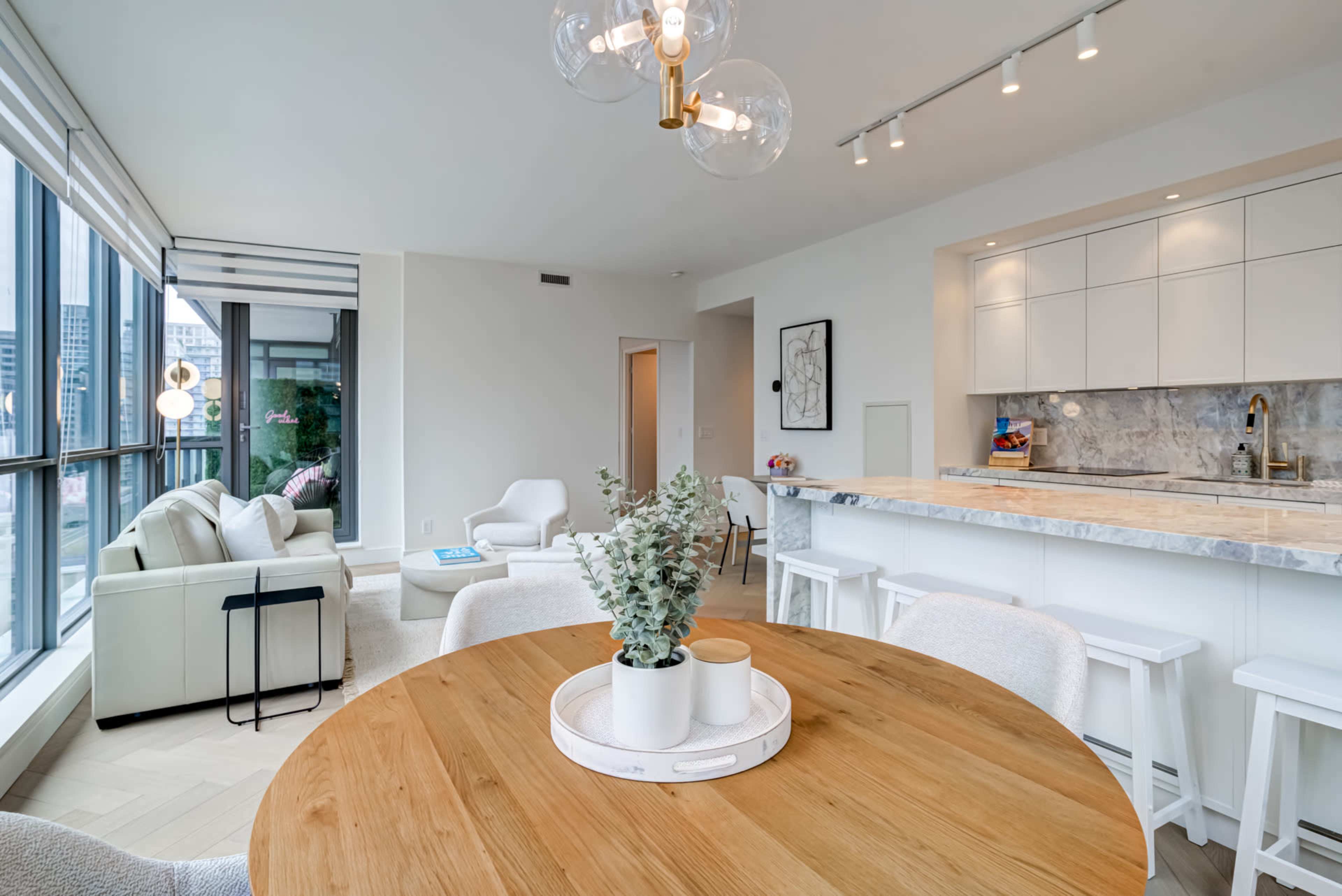 A modern open-concept living and dining area features a round wooden table, white bar stools, and large windows with natural light.