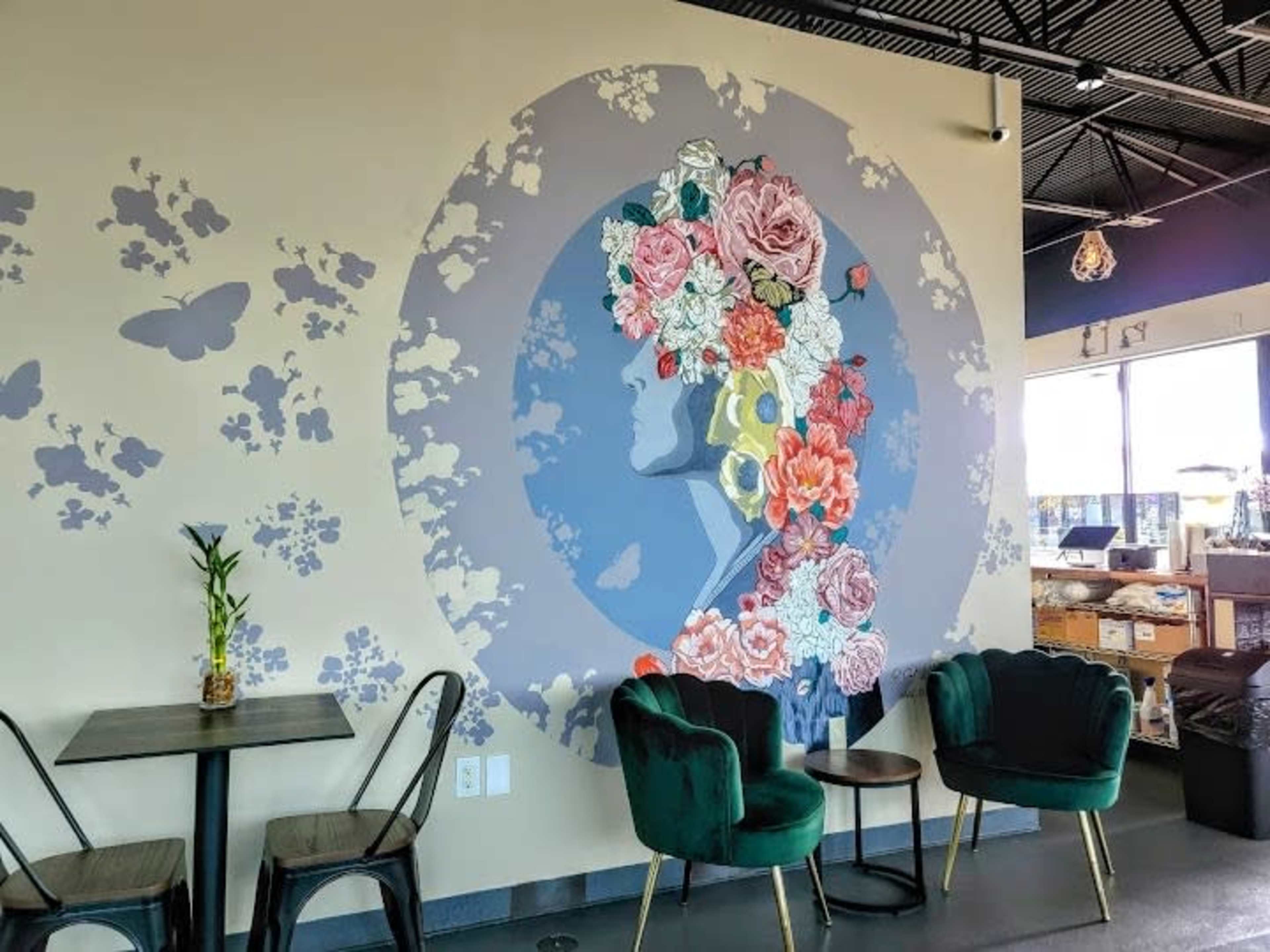 A colorful mural of a floral silhouette on a wall beside two green velvet chairs and a small table in a cafe setting.