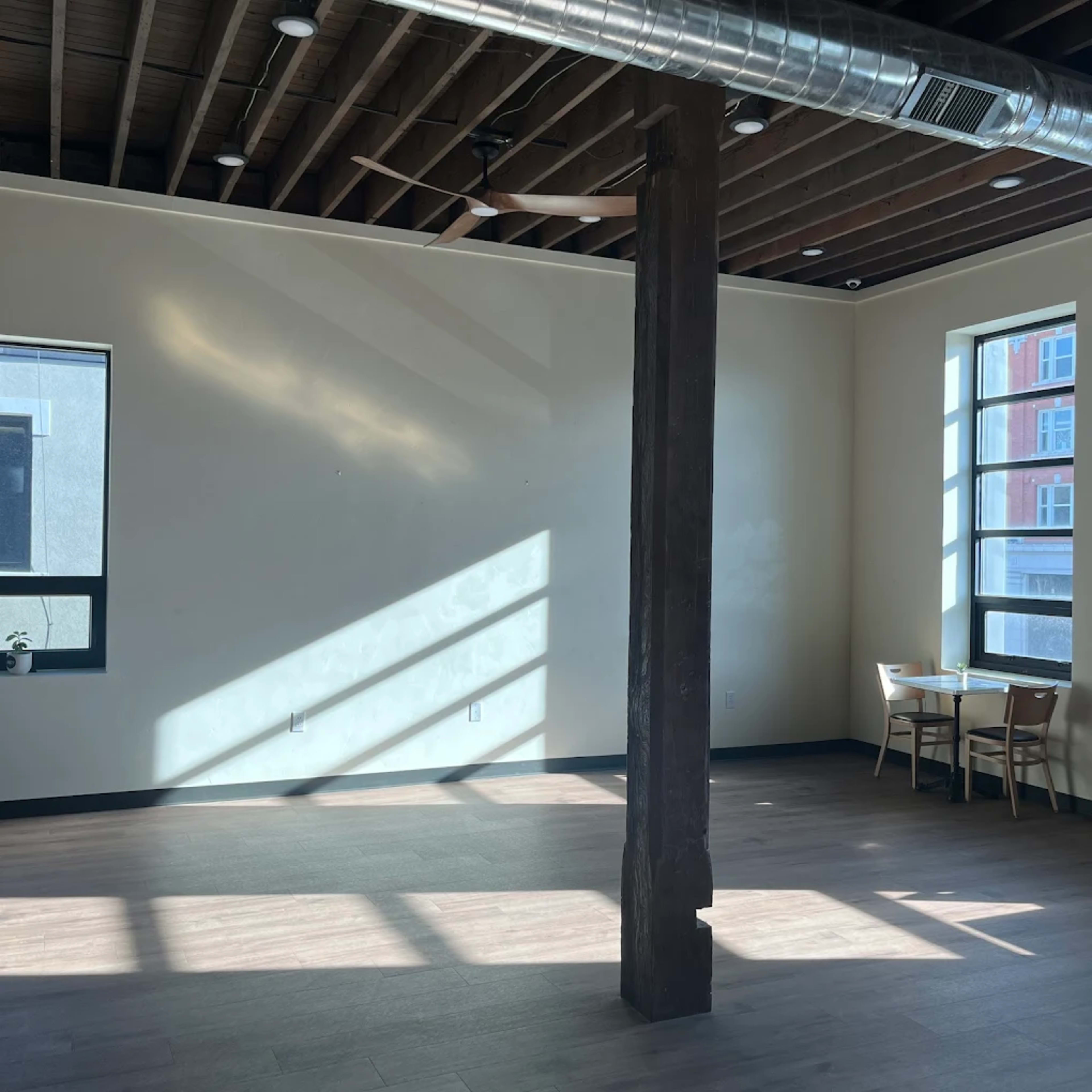 The image shows an empty room with wooden flooring, a large column in the center, and sunlight streaming through the windows.