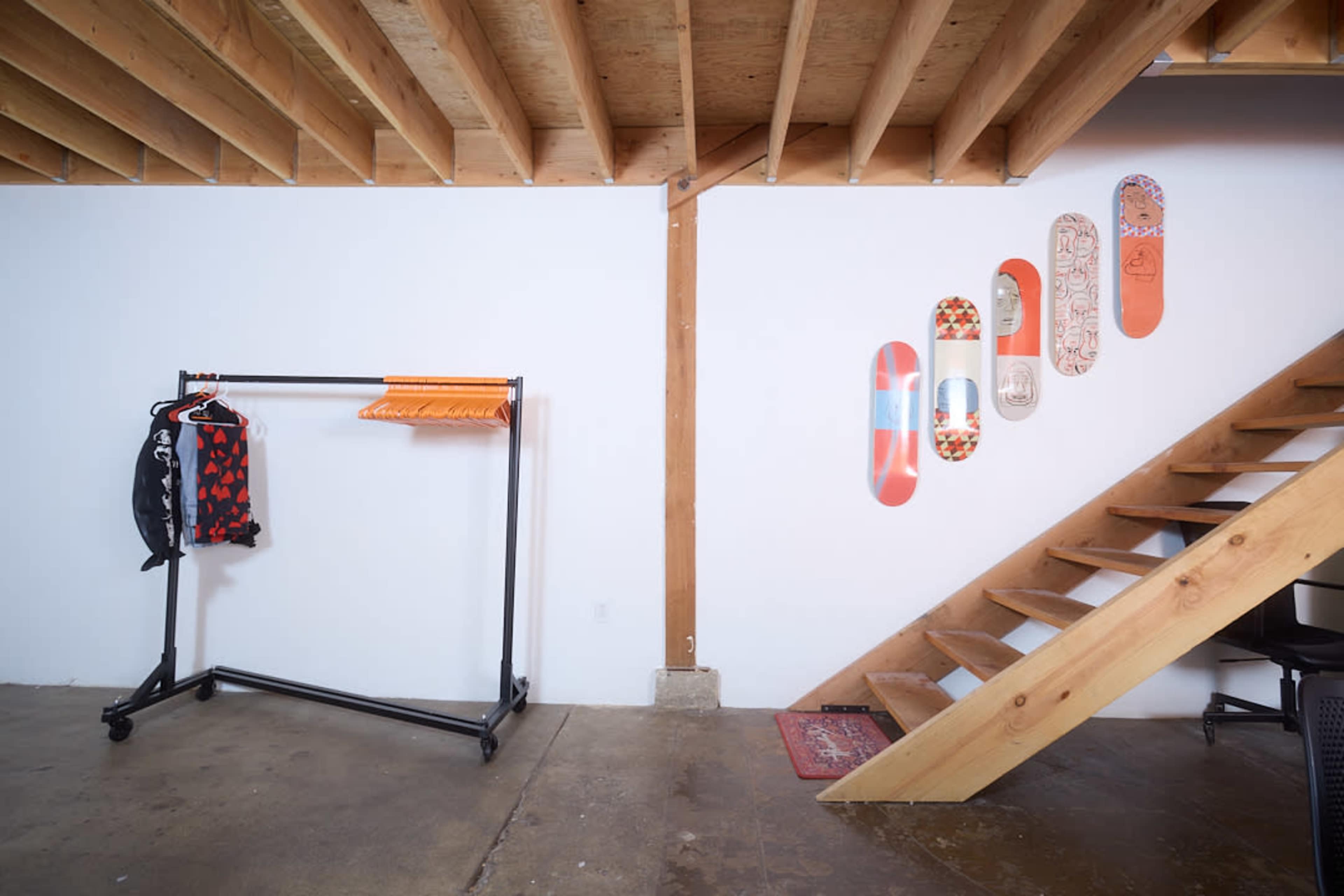 The image shows a modern interior featuring a wooden staircase, a clothing rack with hanging garments, and several decorative skateboards mounted on a white wall.
