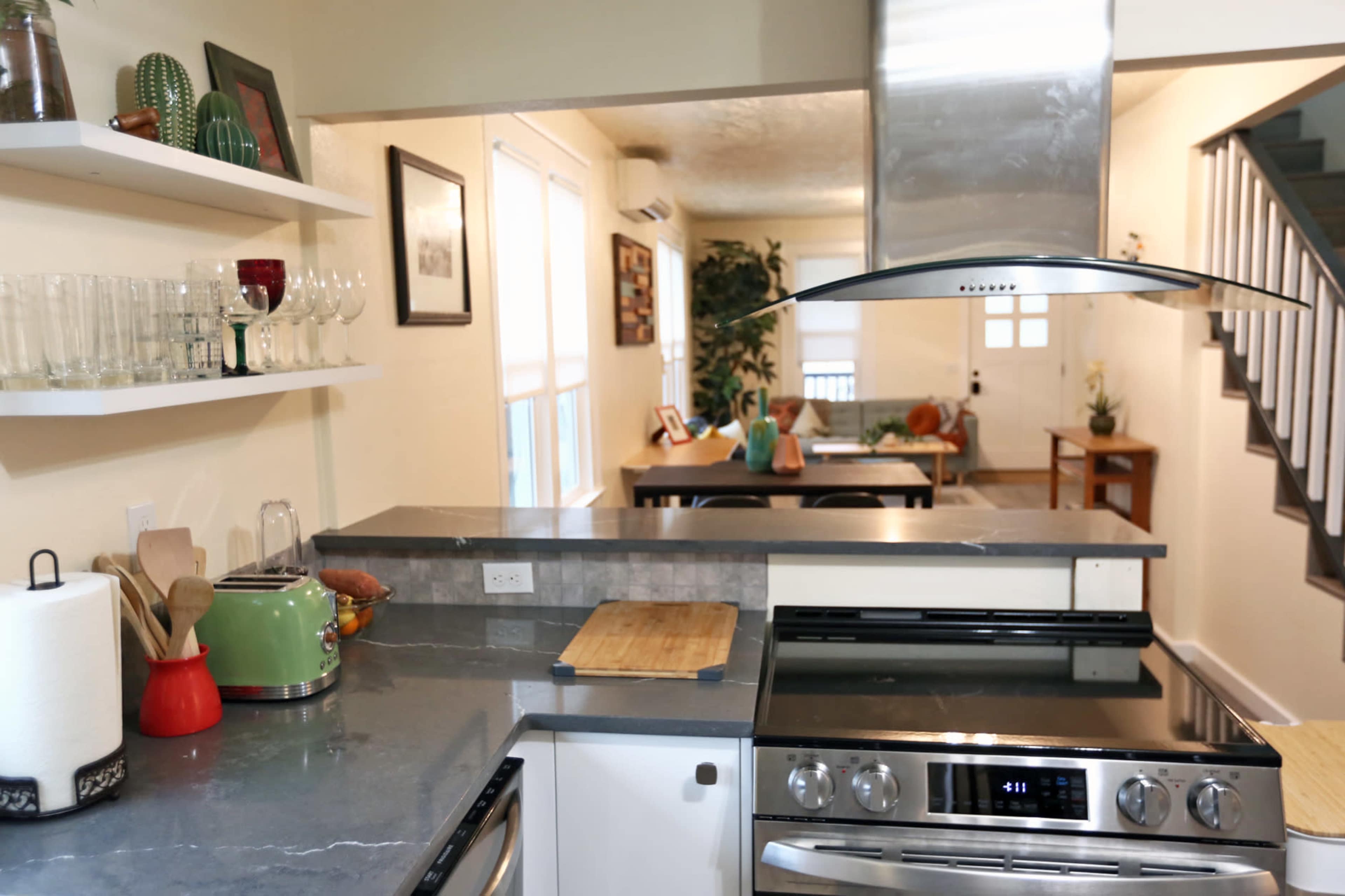 The image shows a modern kitchen with stainless steel appliances, a countertop, and open shelving displaying glassware, leading into a dining area in the background.