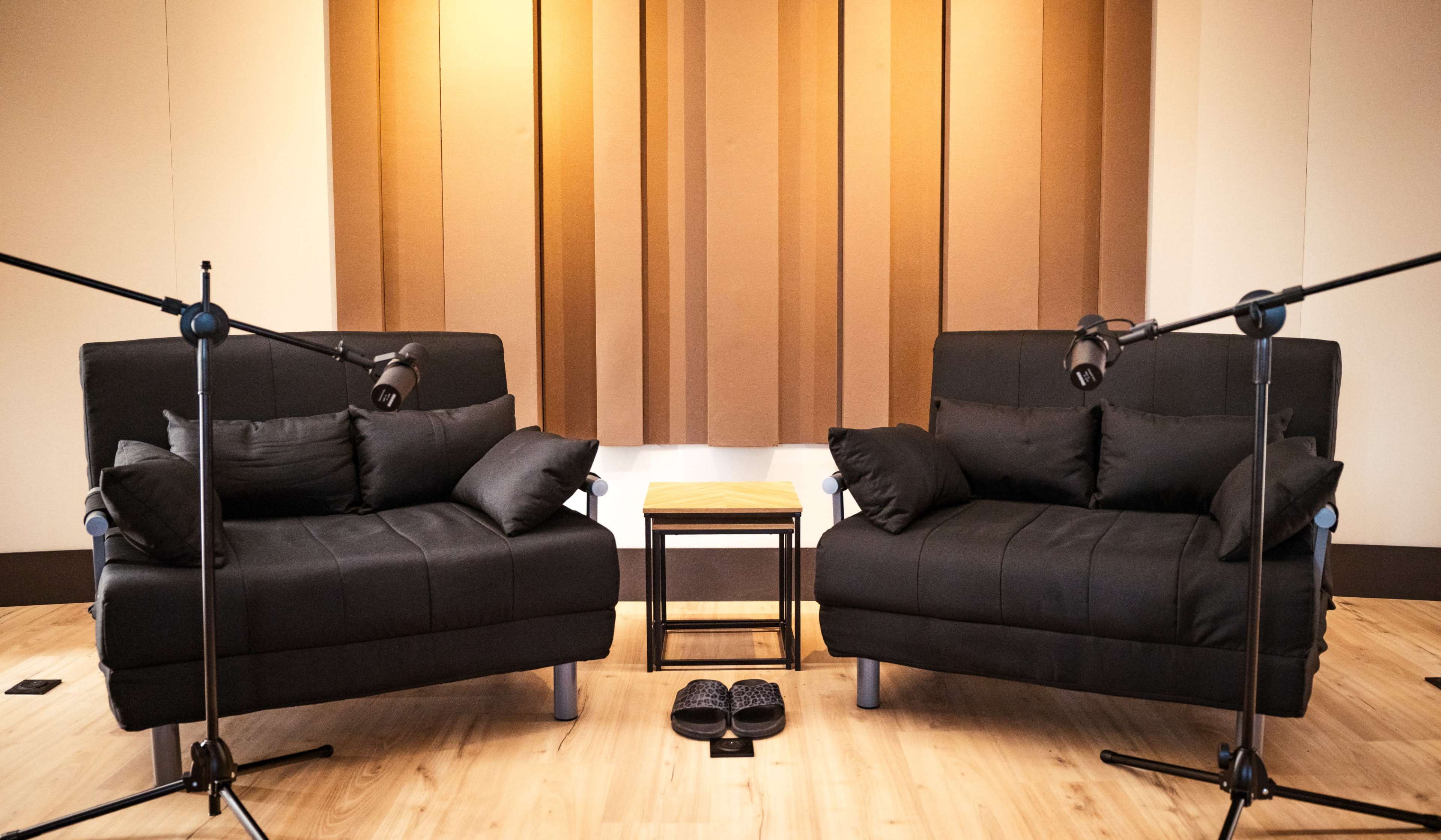 The image shows two black couches facing each other with a small table between them and two adjustable lamps, set against a backdrop of beige soundproofing panels.