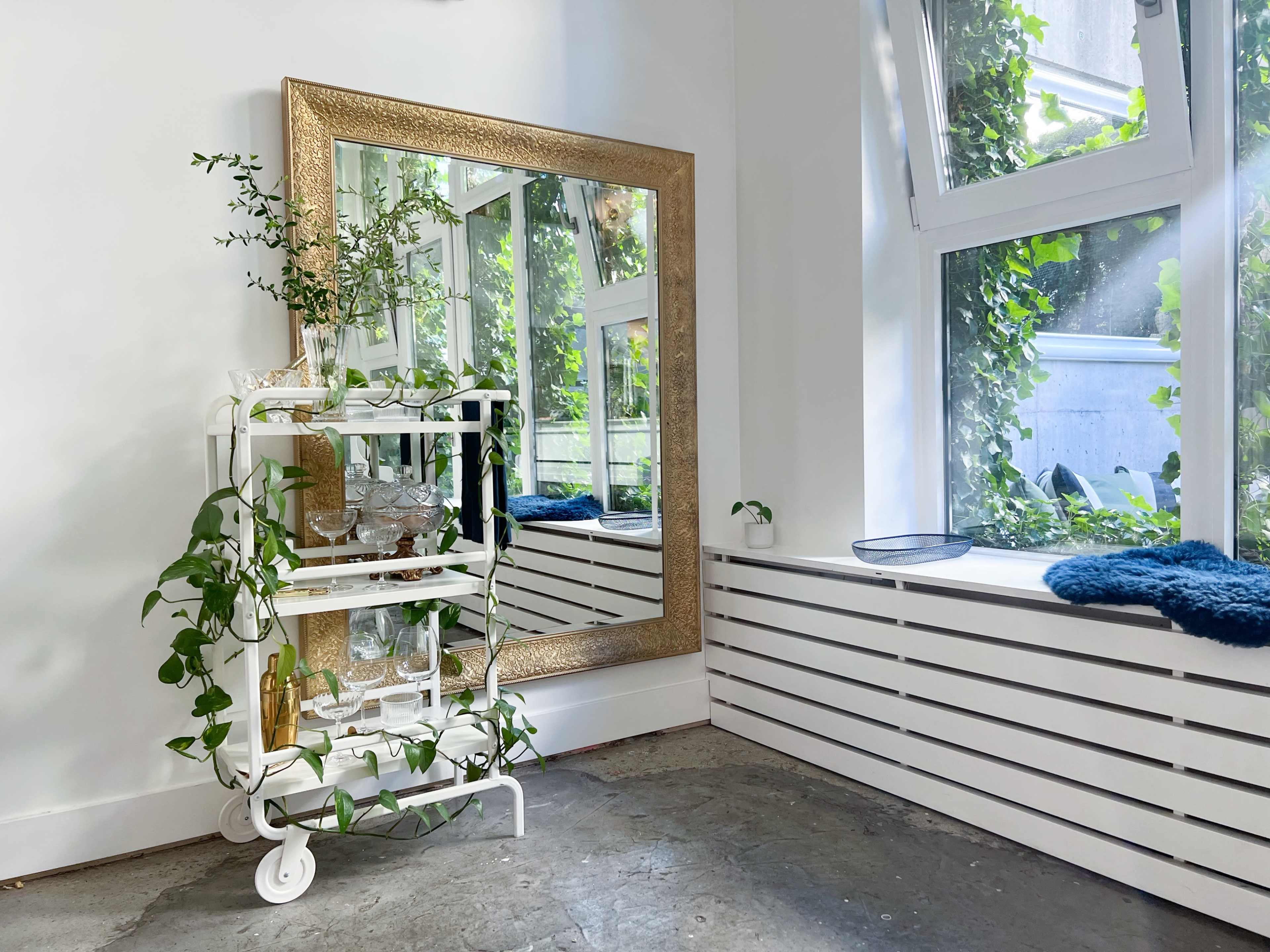 A large gilded mirror and a decorative white shelving unit with plants are positioned beside a window in a bright room.
