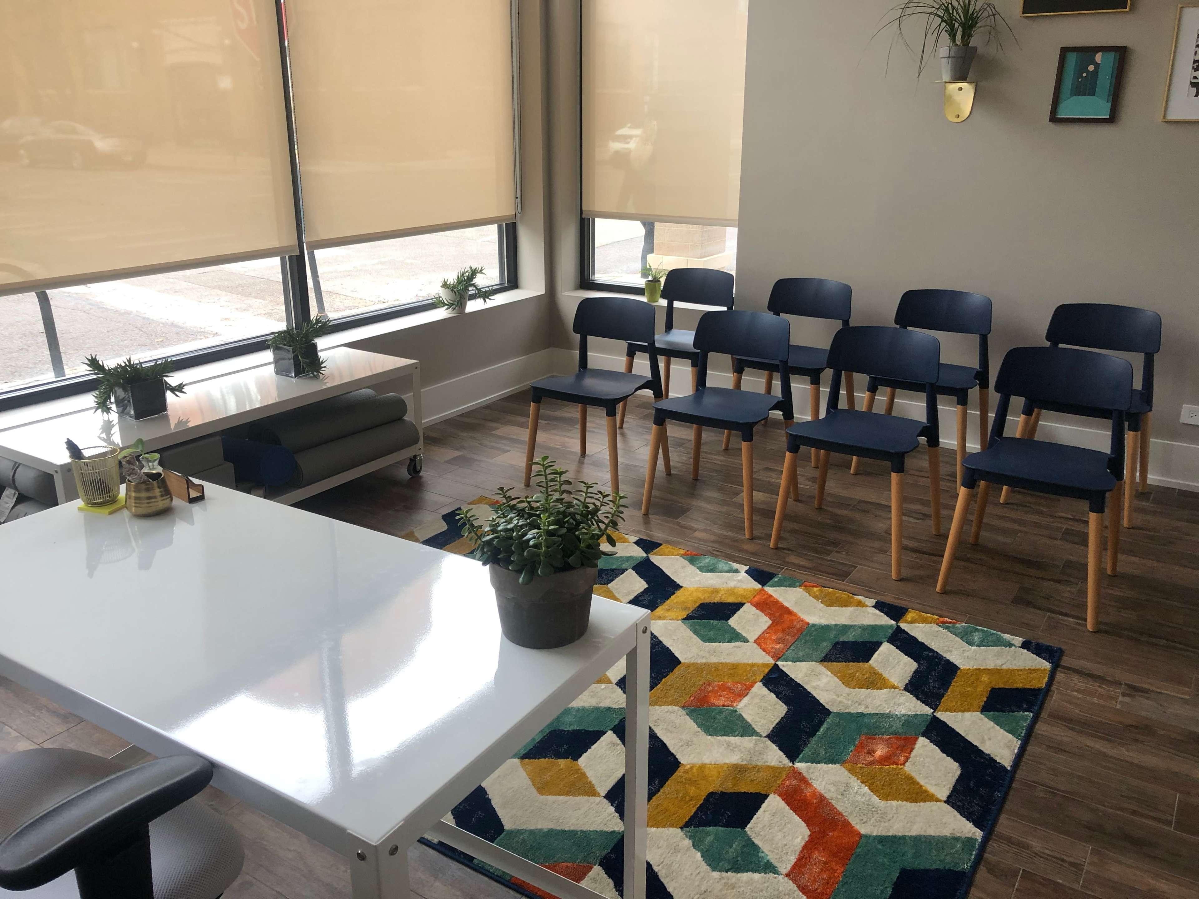 A modern waiting area with a white desk, several blue chairs arranged in rows, and a colorful geometric rug.