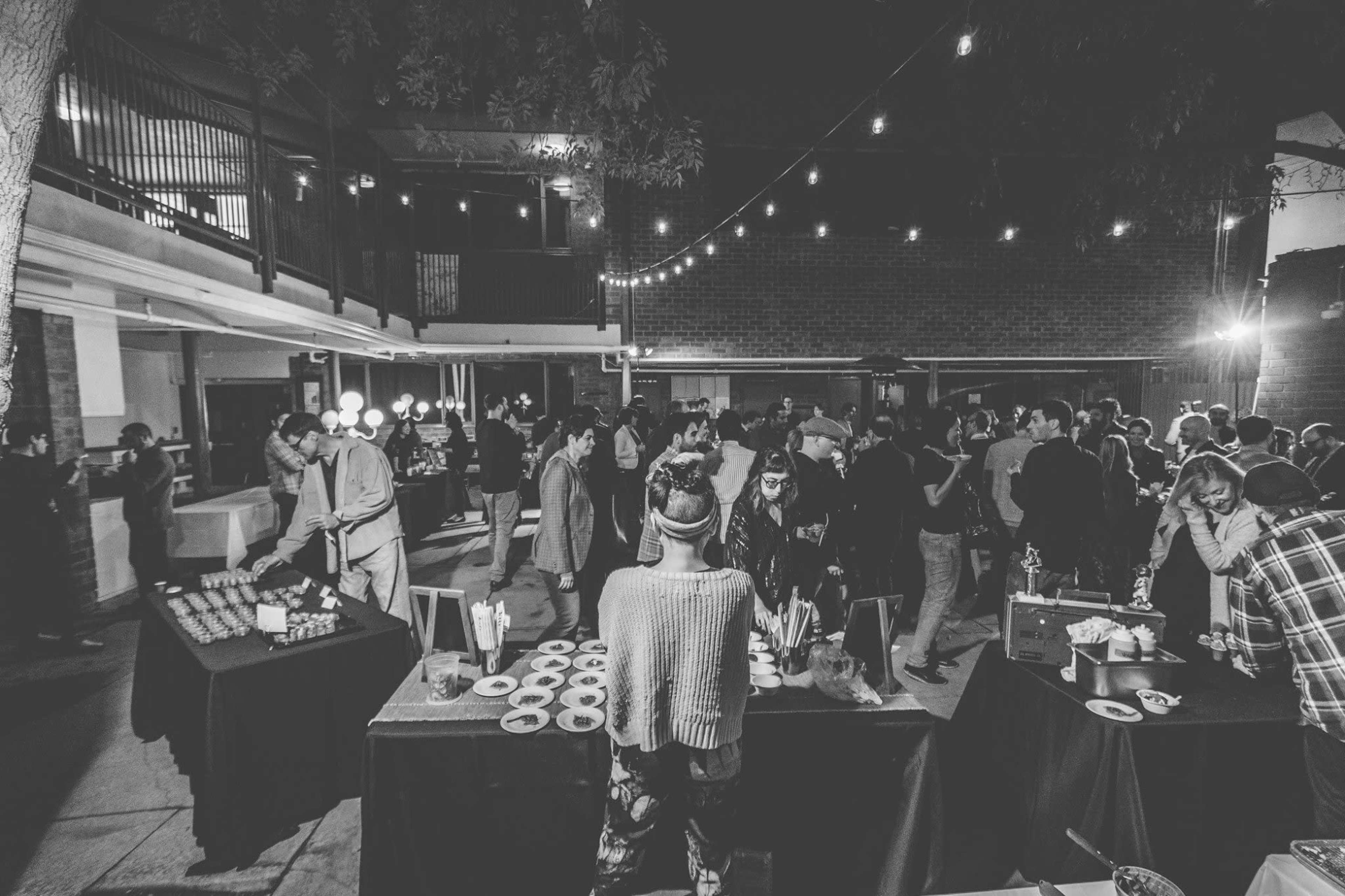 A large group of people mingles at a nighttime outdoor event with string lights illuminating the scene.