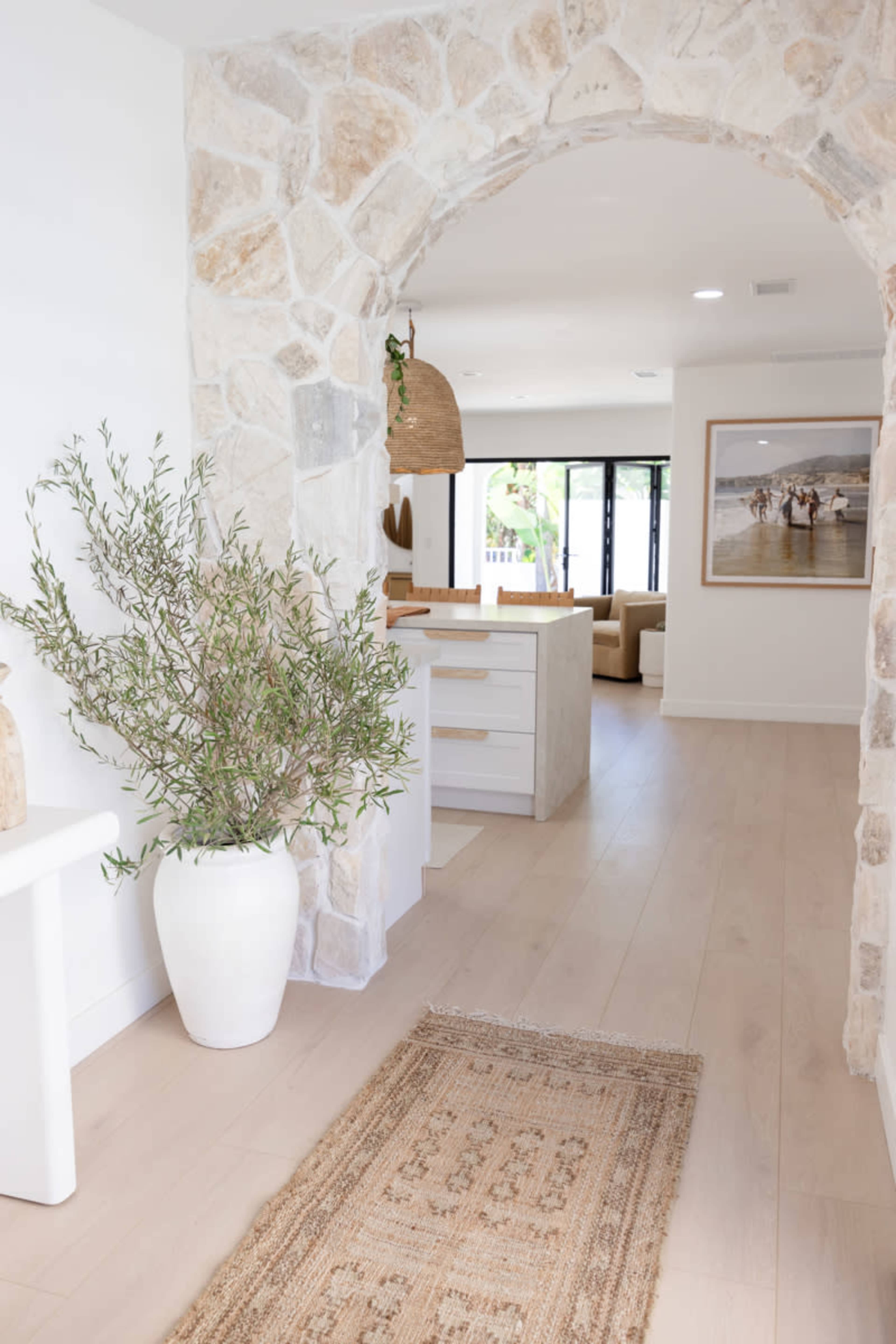 A stone archway frames a light-filled corridor with a modern decor featuring a white vase, a woven rug, and an open living space beyond.