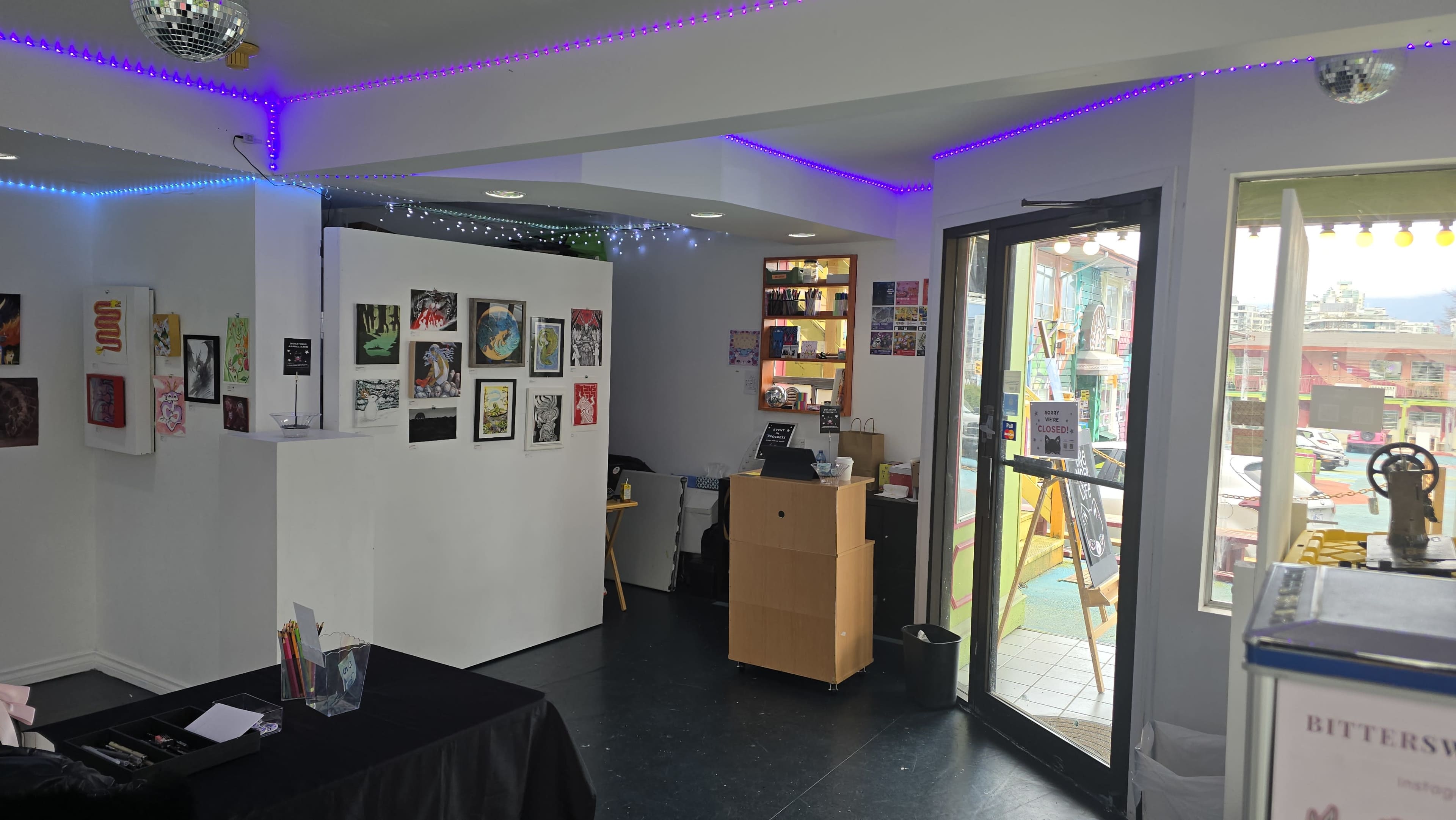 The image shows an art gallery or studio space featuring white walls adorned with colorful artworks, a reception desk, and an open doorway leading to an outdoor area.
