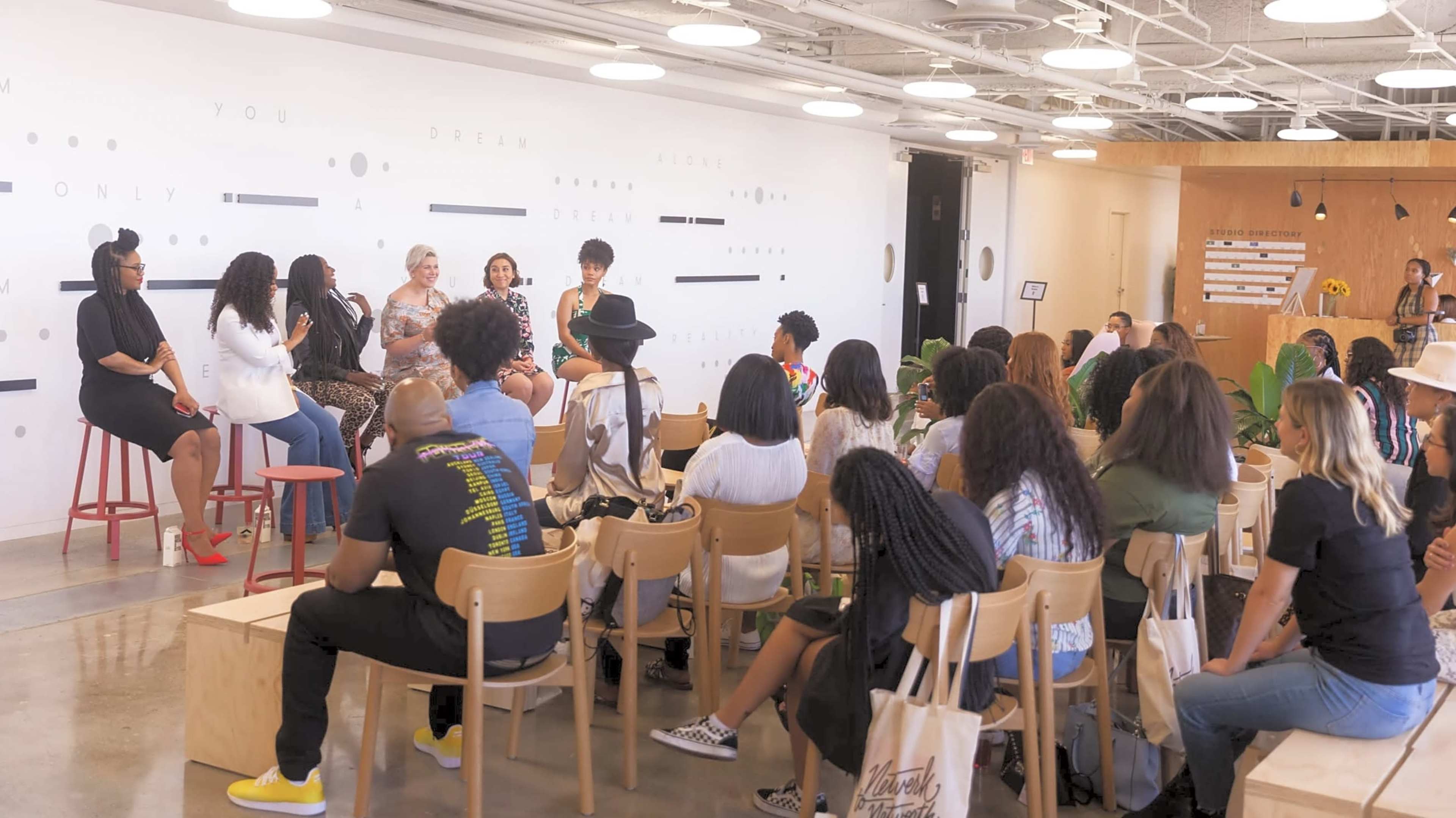 A diverse group of people sits in wooden chairs facing a panel of speakers in a modern event space with a minimalist decor.