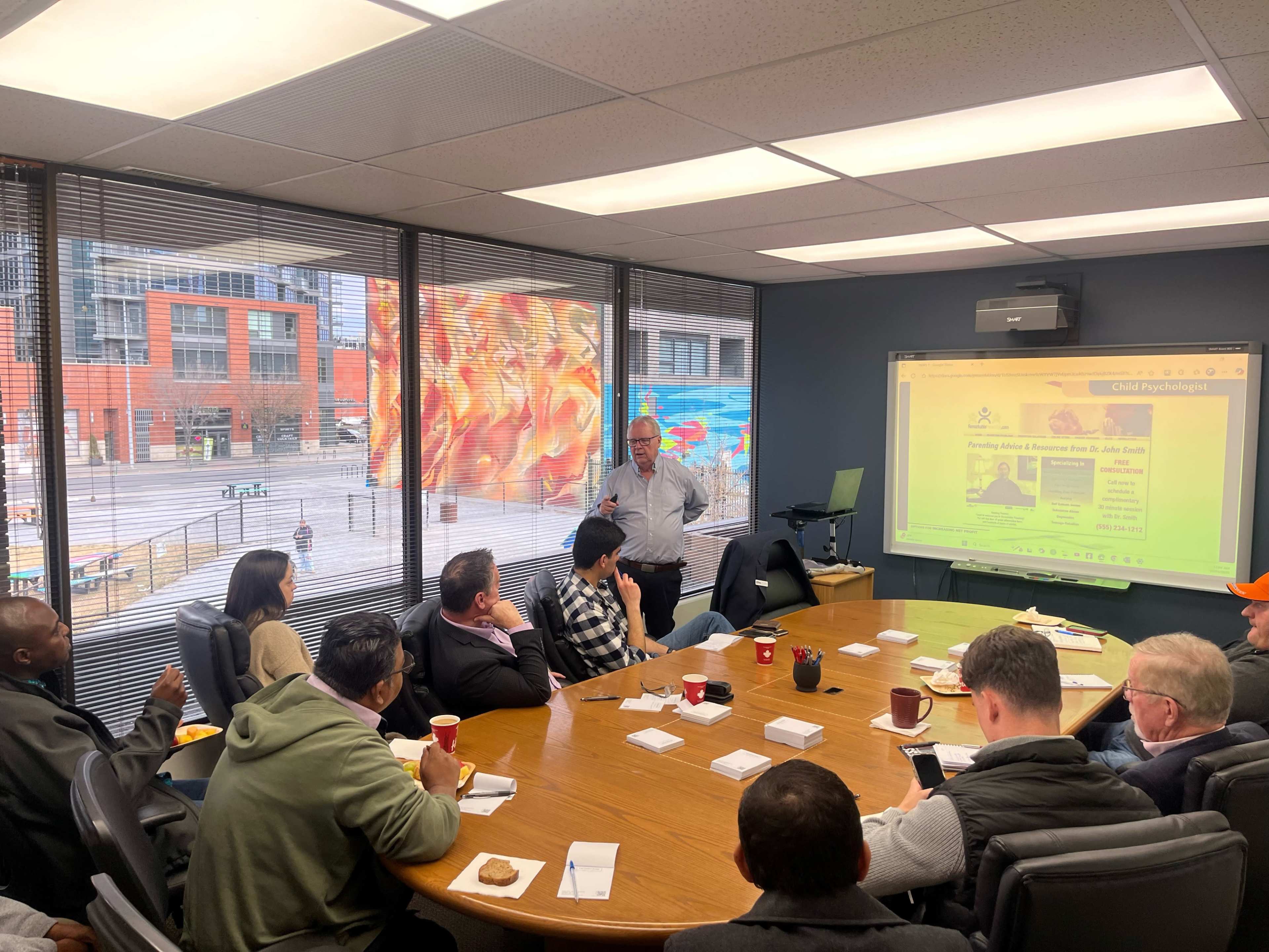 A group of people is seated around a conference table, while a man stands in front of a screen displaying a presentation in a brightly lit meeting room with large windows.